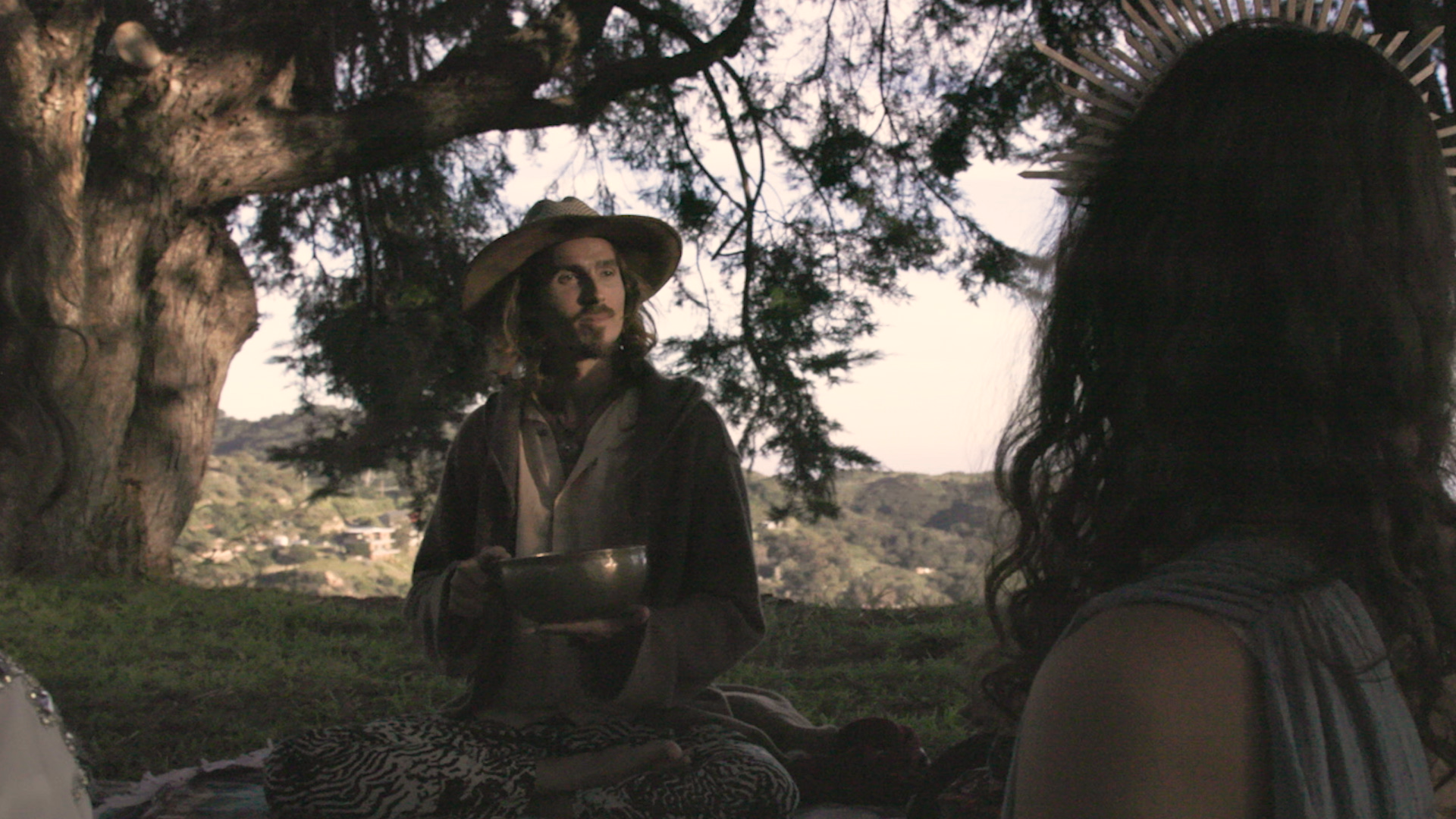 Dylan Townsend with long hair and a mustache wearing a wide-brimmed hat and holding a clipboard, talking to a woman with long dark hair and a straw hat in a shaded outdoor setting with trees and a hilly landscape in the background.
