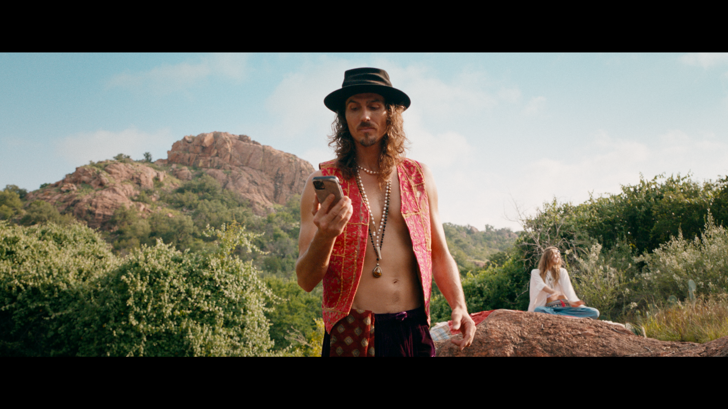 Dylan Townsend with long curly hair wearing a black hat, a red patterned vest, and layered necklaces, looking at his phone in a natural outdoor setting with greenery and a rocky hill in the background.
