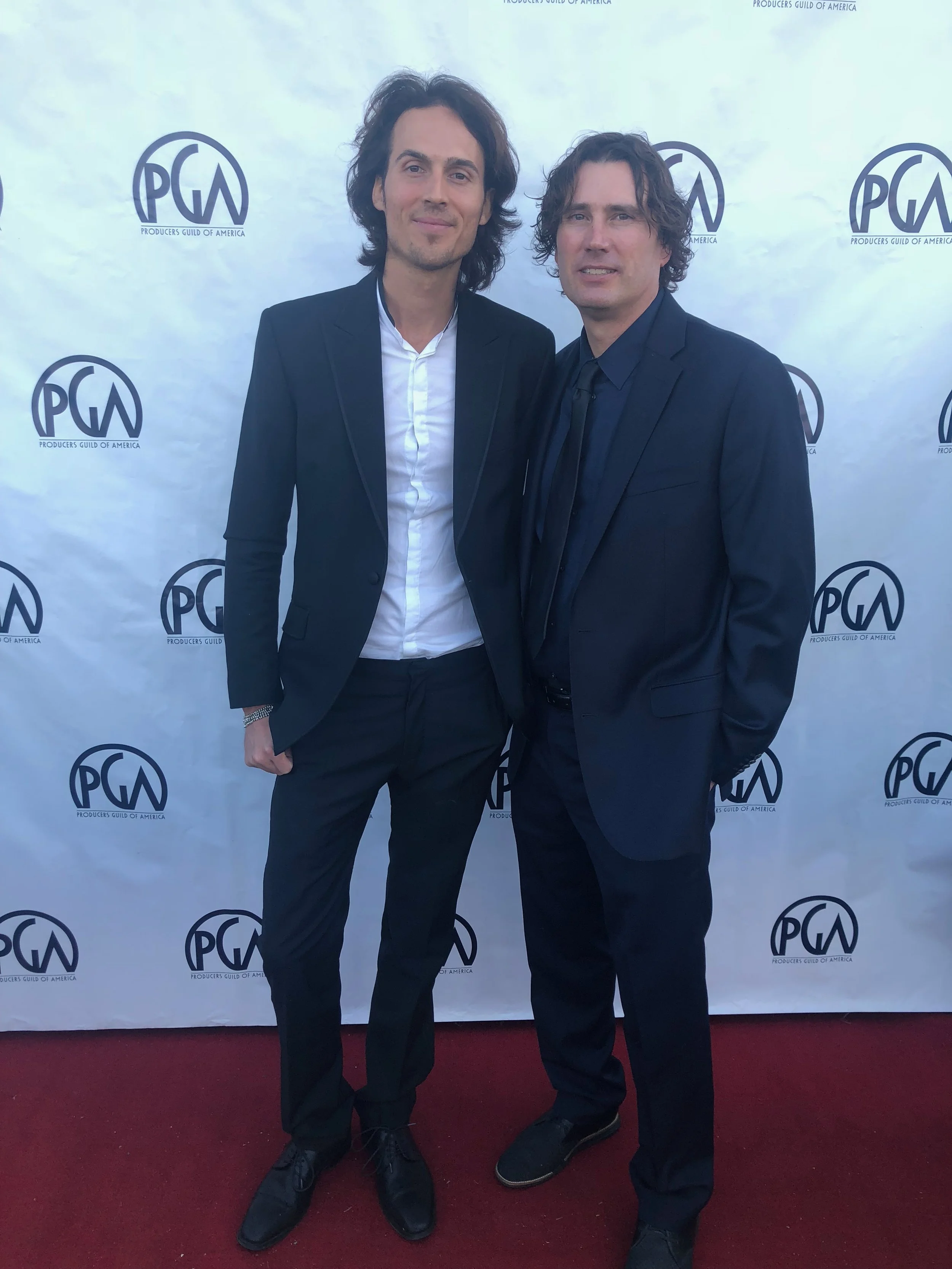 Dylan Townsend. PGA. Producers guild. Two men in black suits standing on a maroon carpet in front of a white backdrop with the PGW logo, posing for a photo at an event.