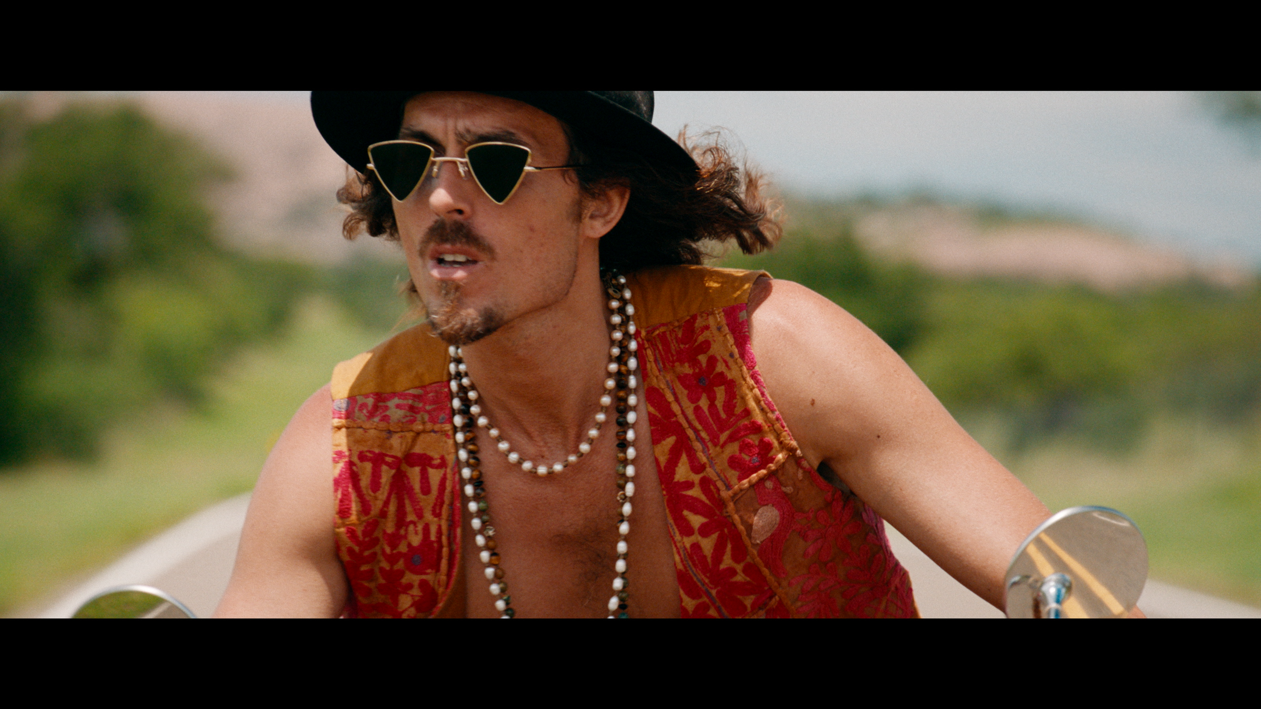 A man riding a motorcycle outdoors, wearing sunglasses, a black hat, a patterned sleeveless shirt, and multiple necklaces, with greenery and a blue sky in the background.