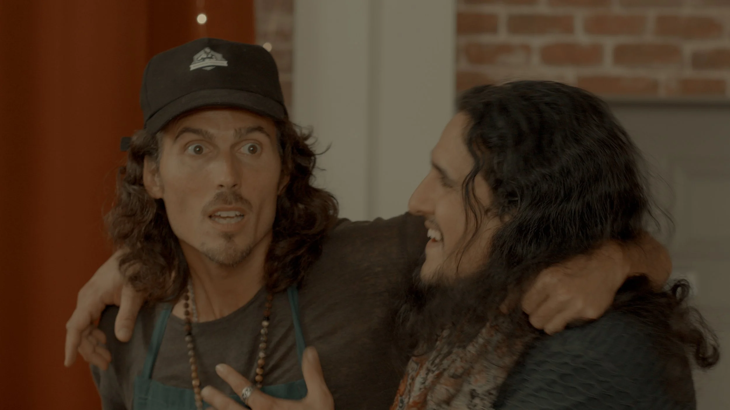 Dylan Townsend and a man with long hair and beards smiling and hugging in a room with a brick wall background.