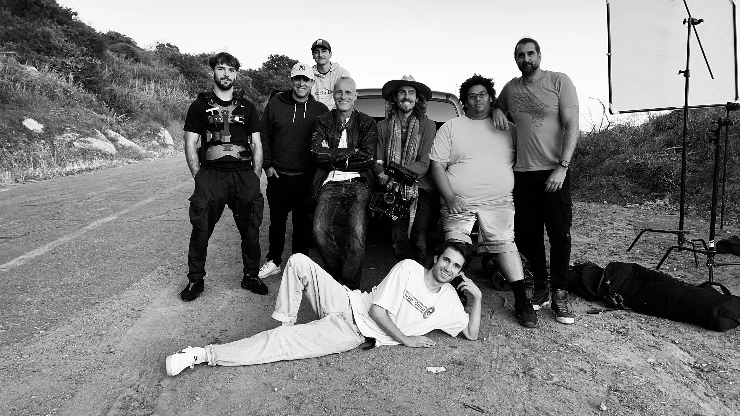 Dylan Townsend BTS with Timothy V. Murphy posing outdoors with equipment, some standing and one lying on the ground, near a car and for photography setup, hillside in the background, black and white photo.