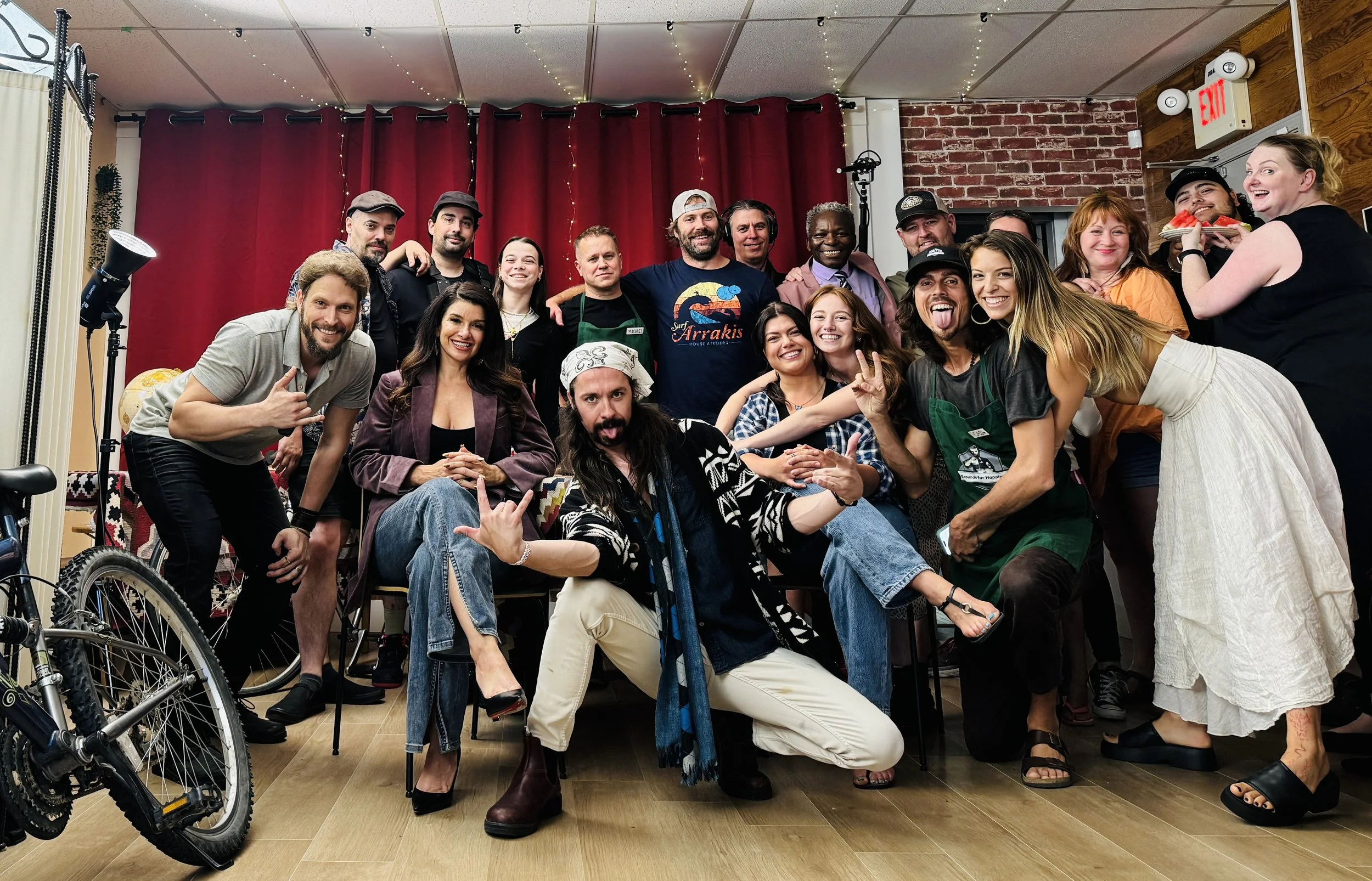 Under The Influence Tv show wrap. Dylan Townsend. Group of diverse people smiling and posing inside a room with red curtains, brick wall, and string lights, some making playful gestures, and one person holding a slice of watermelon.