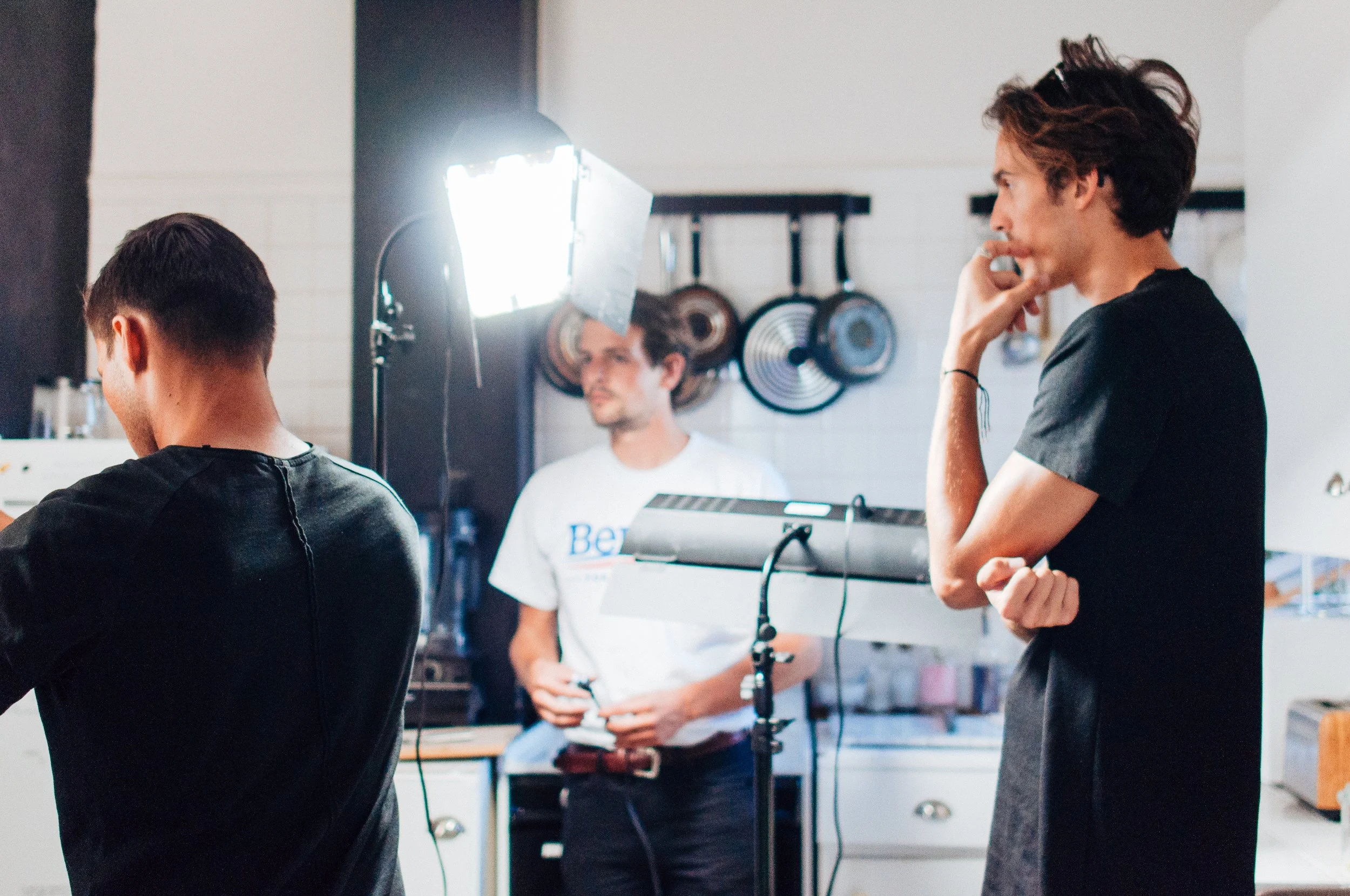 Dylan Townsend onset directing with film equipment, one standing and two seated, with a bright light on a stand. The person on the right is smiling and resting his chin on his hand, while the other two are focused on something out of frame.