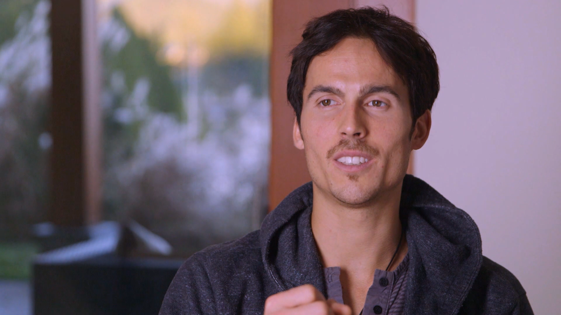Dylan Townsend on Tv show 'Exiles Vancouver'. A man with short dark hair and light skin is smiling slightly, looking off to the side. He is wearing a dark gray hoodie over a gray button-up shirt.