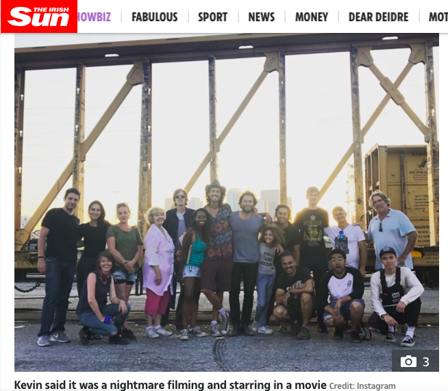The Sun article with Dylan Townsend & Kevin Ryan onset. A Group of people posing together outdoors in front of a train car and large metal structures during sunset.