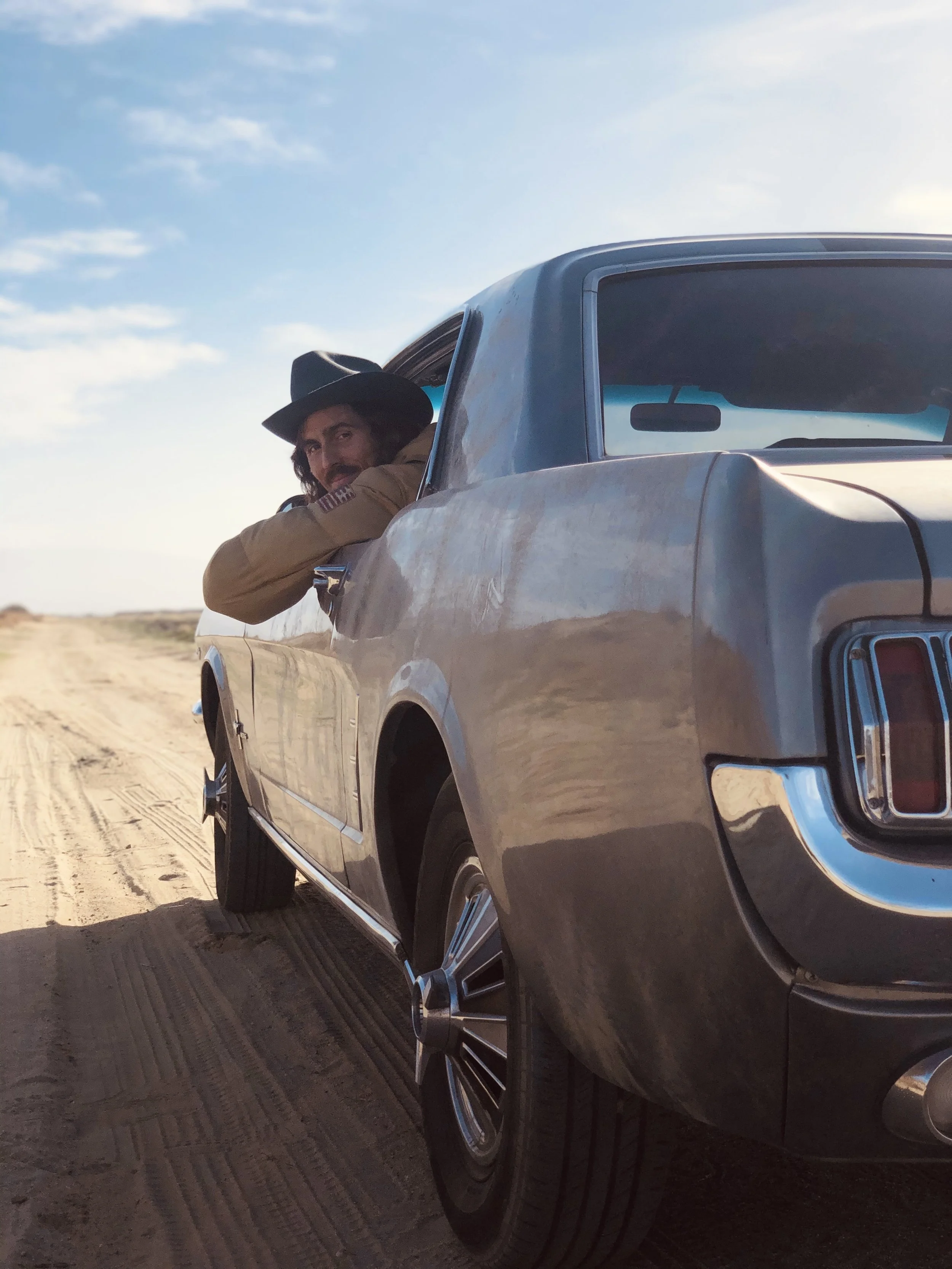 Dylan Townsend on set BAD SUNS with long hair and a beard wearing a cowboy hat leaning out of the window of a vintage pickup truck on a dirt road in a desert landscape under a blue sky with some clouds.