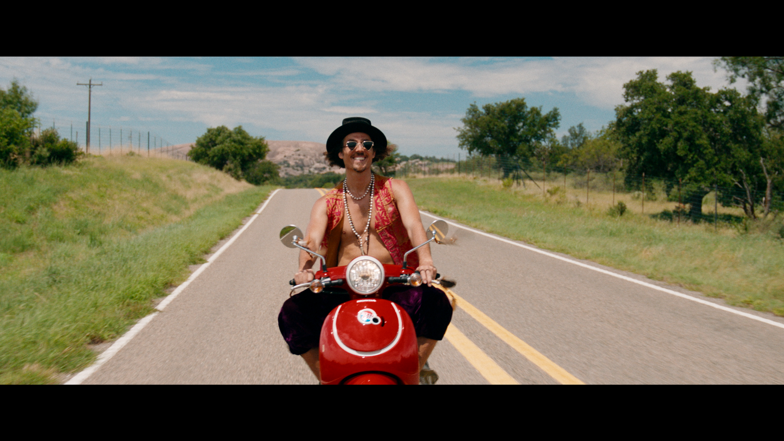 Dylan Townsend wearing sunglasses, a black hat, a patterned vest, and multiple necklaces rides a red scooter down a rural road with green trees on either side under a partly cloudy sky.
