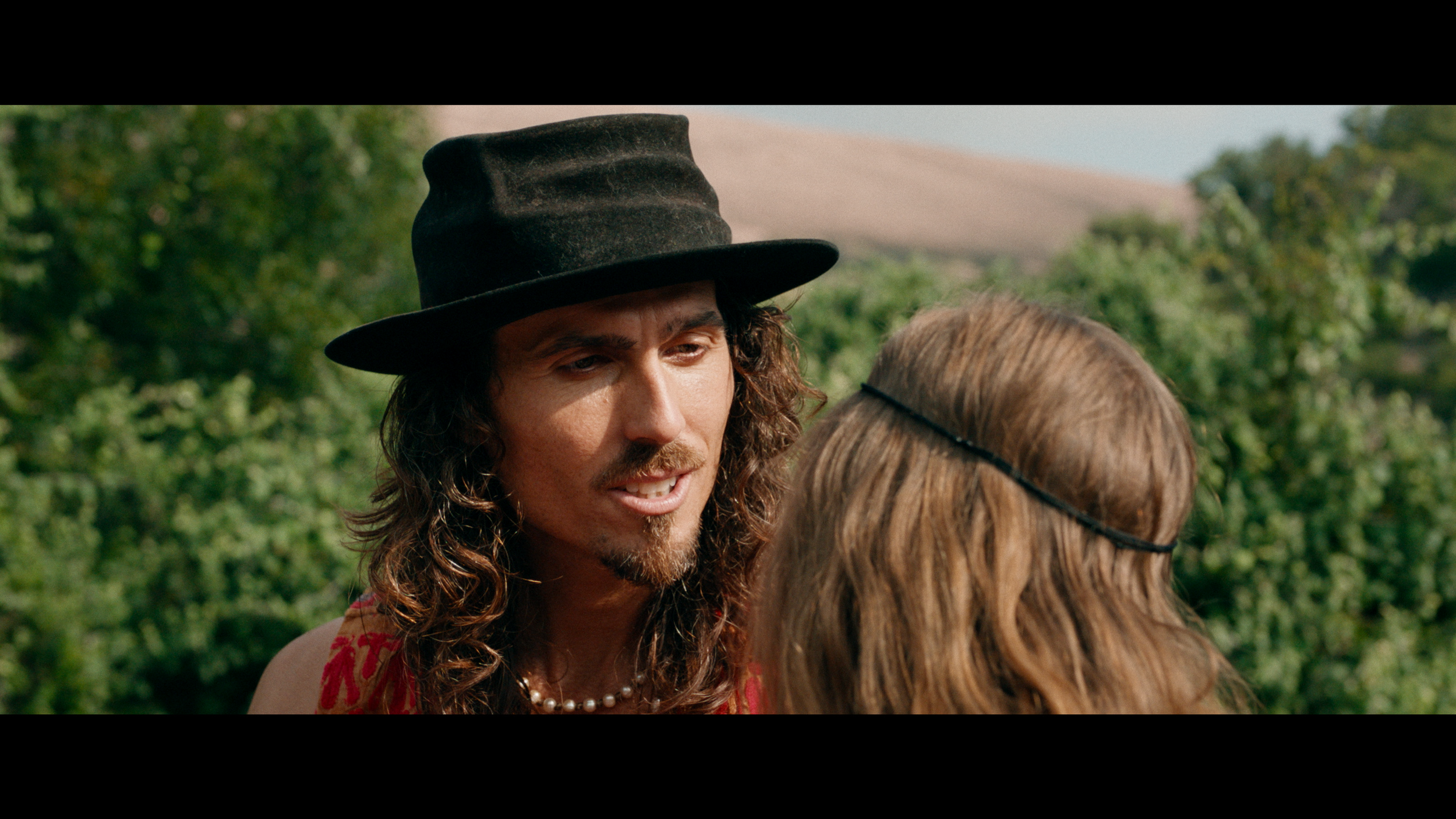 Dylan Townsend with long curly hair, wearing a black hat and red clothing, is talking to a woman with light brown hair who is wearing a headband, outdoors with green trees in the background in feature film ' The Academy of Everything is Possible.'