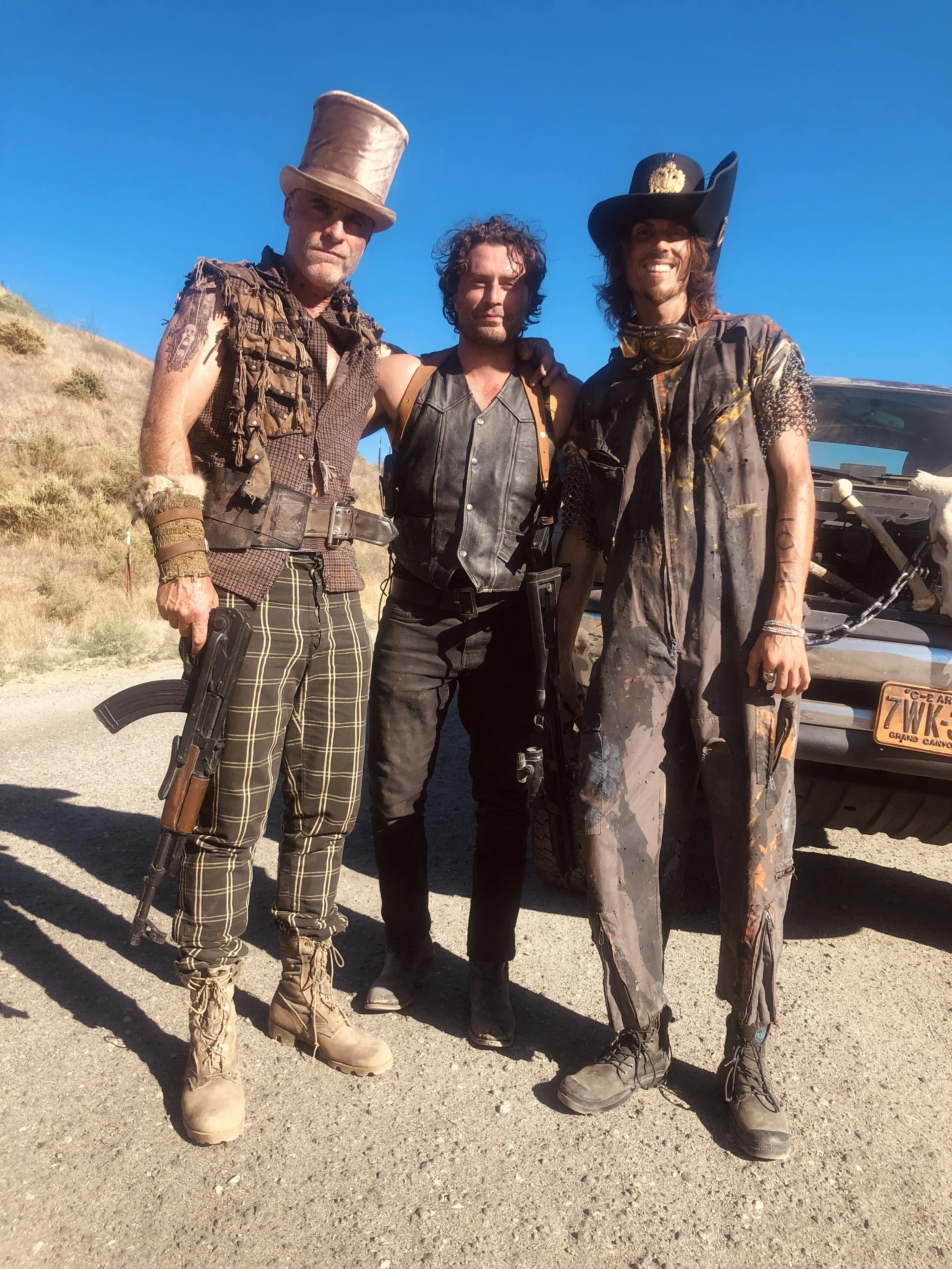 Dylan Townsend, Timothy V. Murphy & Kevin Ryan on set BAD SUSNS. Three individuals dressed in post-apocalyptic costumes, standing outdoors with a vehicle in the background and a clear blue sky.