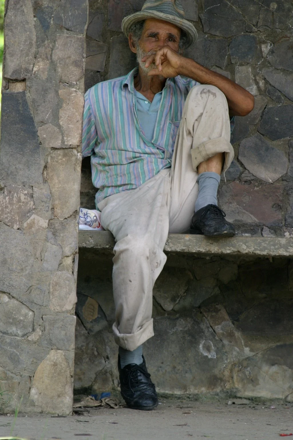An elderly man sitting on a stone ledge, wearing a hat, striped shirt, beige pants, and black shoes, leaning against a stone wall, with his hand covering his mouth