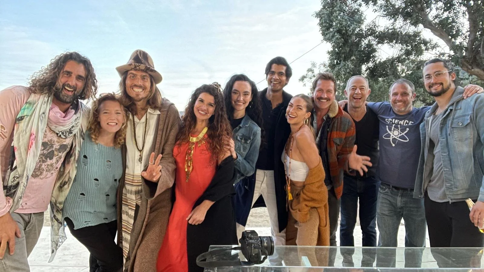 Dylan Townsend, Kirsten Sheridan onset and eleven diverse people smiling and posing outdoors with trees and a cloudy sky in the background. There is a camera on a table in the foreground.