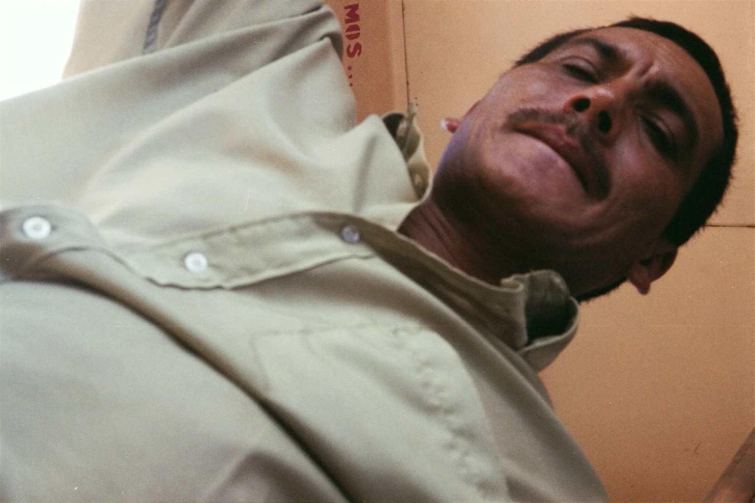 A man with short dark hair and a mustache, wearing a beige button-up shirt, smiling slightly in an indoor setting with beige walls.