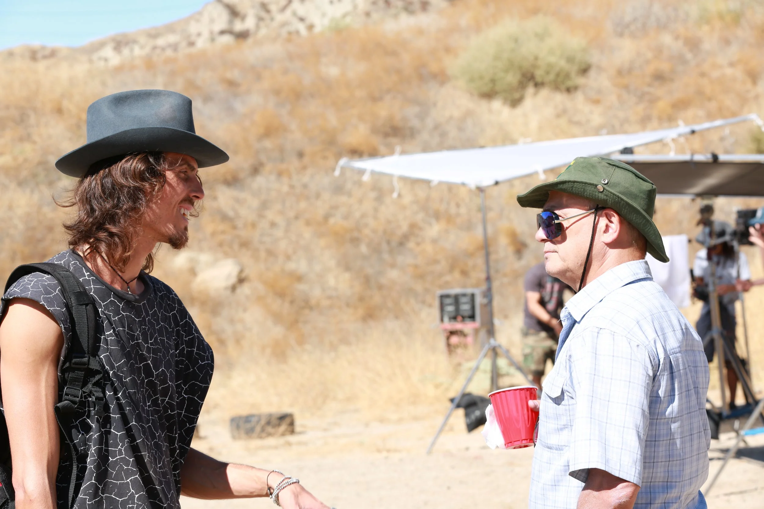 Dylan Townsend on set BAD SUNS facing another man engaged in conversation.  The background shows dry, hilly terrain and medical or filming equipment, with other people and equipment visible in the distance.