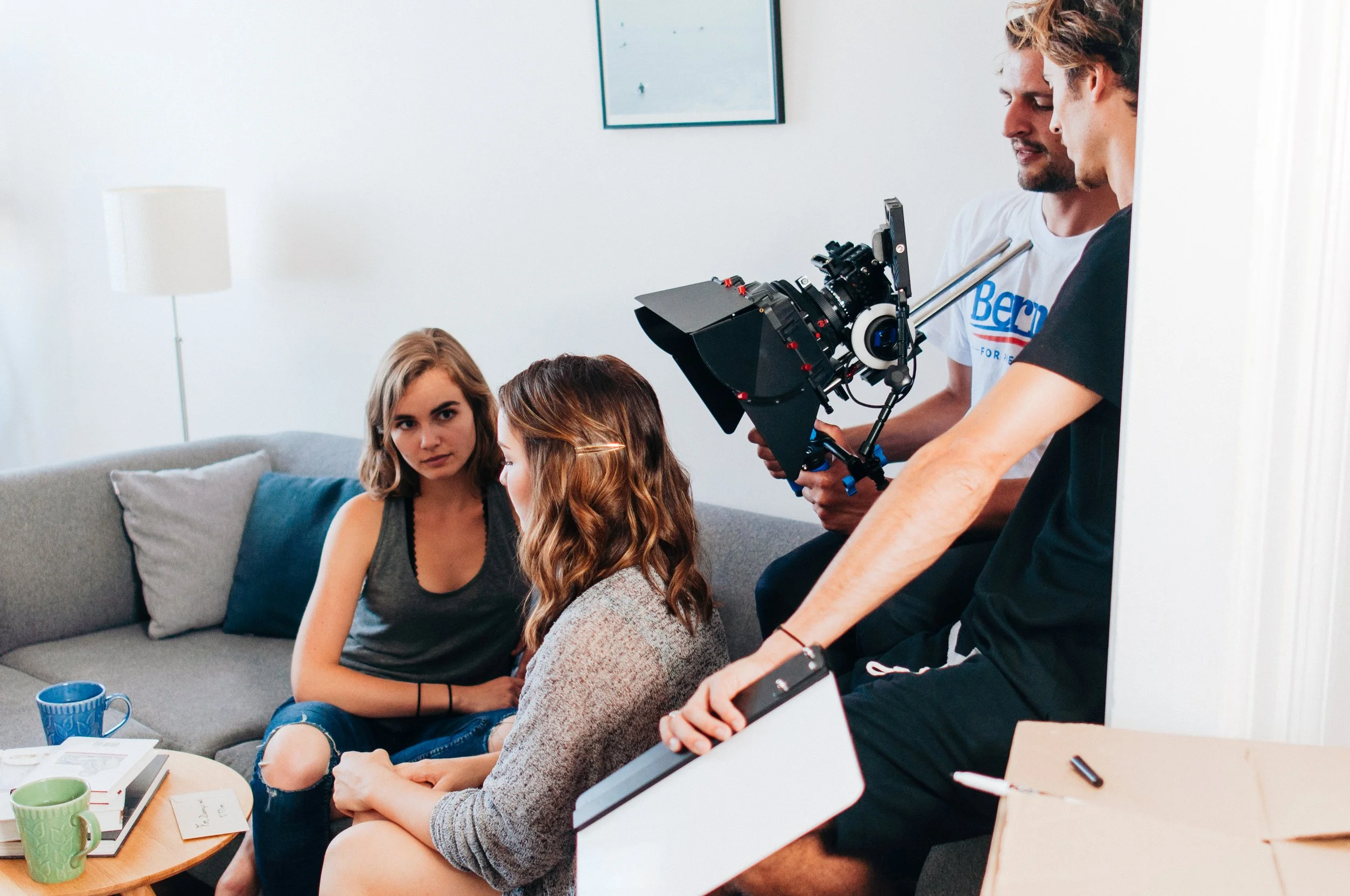 Three young women participating in a filmed conversation with Dylan Townsend directing . The women have long hair and casual clothing, and the scene takes place in a cozy, well-lit room with wall art and a table with cups and papers.