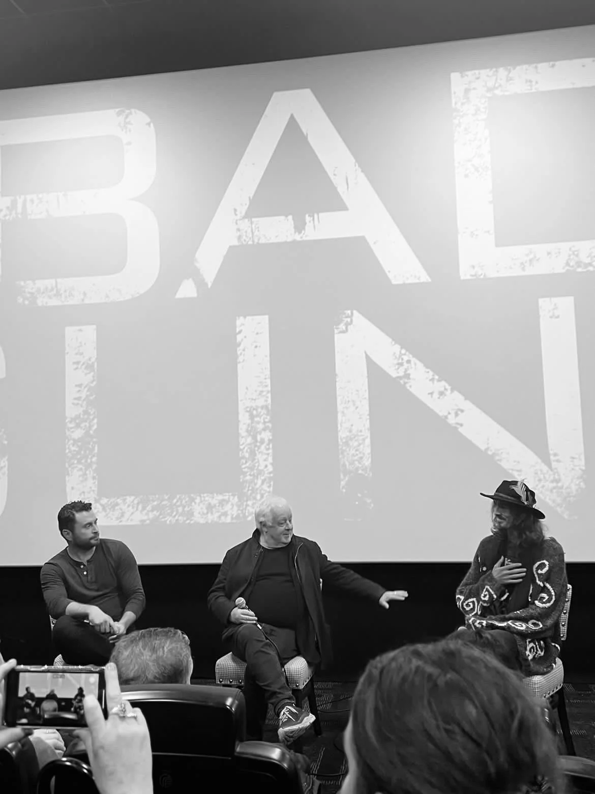 Dylan Townsend, Jim Sheridan & Kevin Ryan are sitting in chairs, for a Q & A, after a screening of BAD SUNS. With a large screen behind them displaying the word in bold, distressed font. Audience members are visible in front.