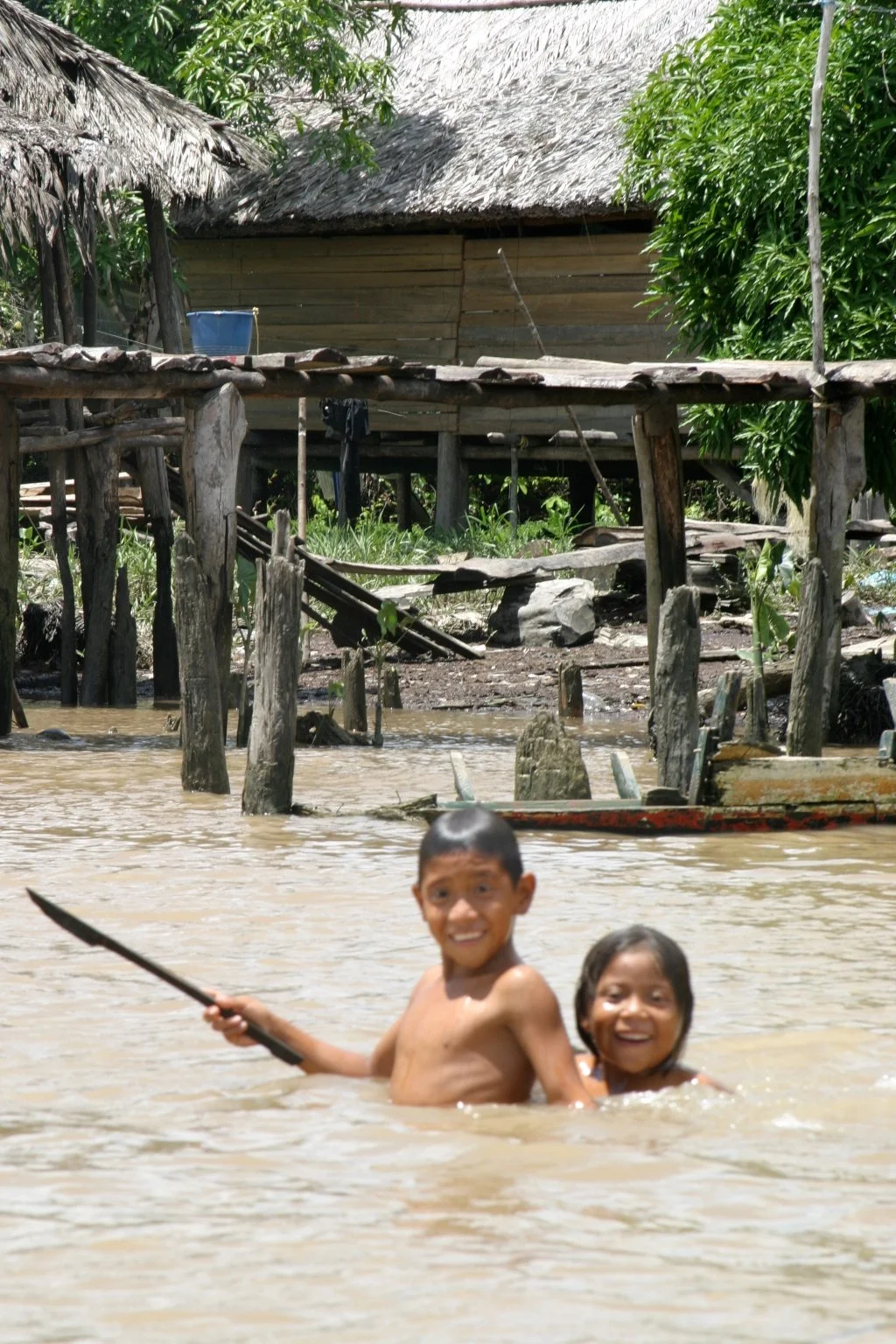 Two children swimming in muddy water near a stilted wooden house and lush green trees in a rural setting.