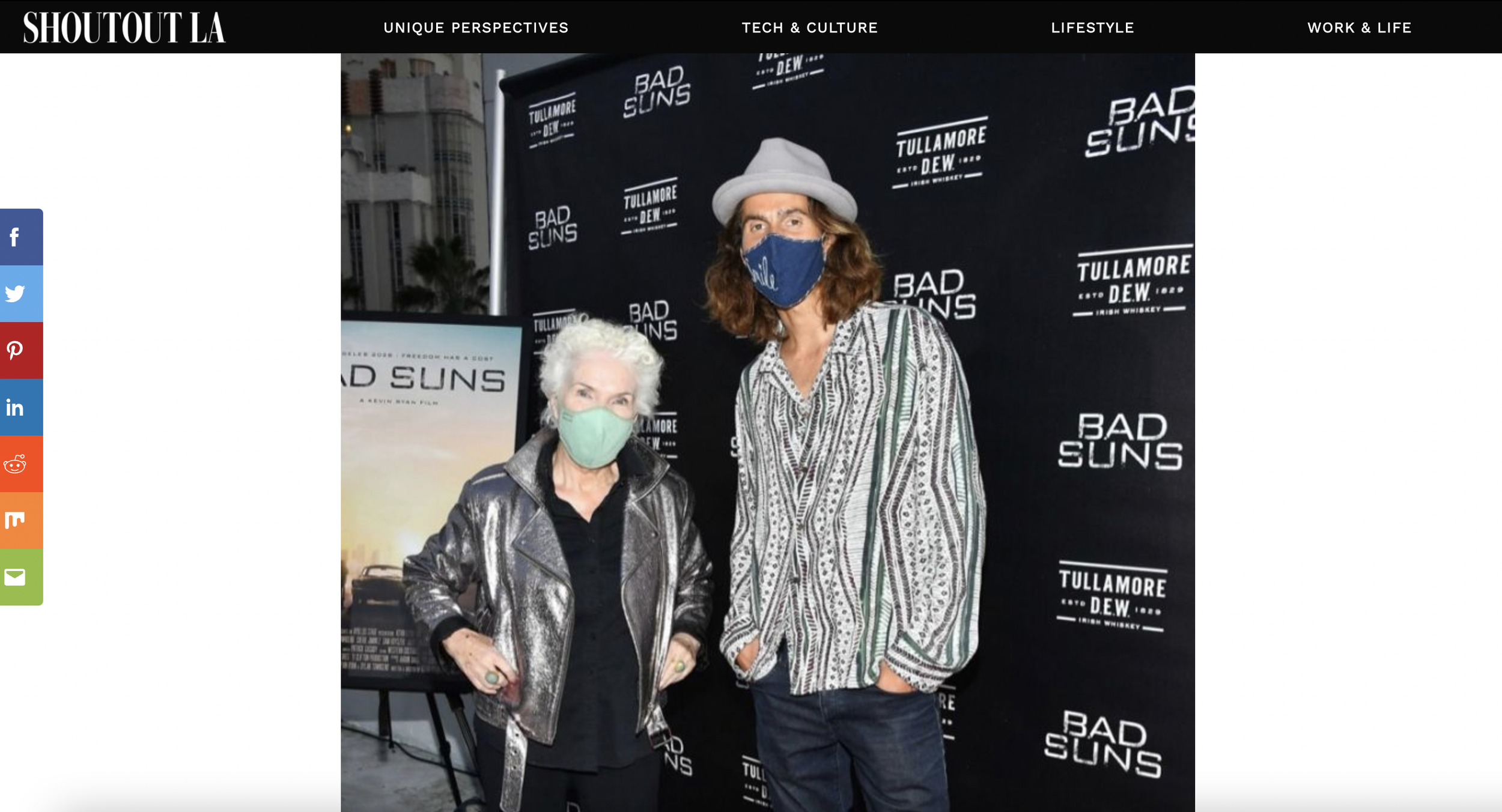 A Man and women. Dylan Townsend and Fionnula Flanagan. wearing face masks standing in front of a backdrop that has the text "BAD SUNS" and "TULLAMORE D.E.W." The woman on the left has white hair, a metallic jacket, and a light green mask. 