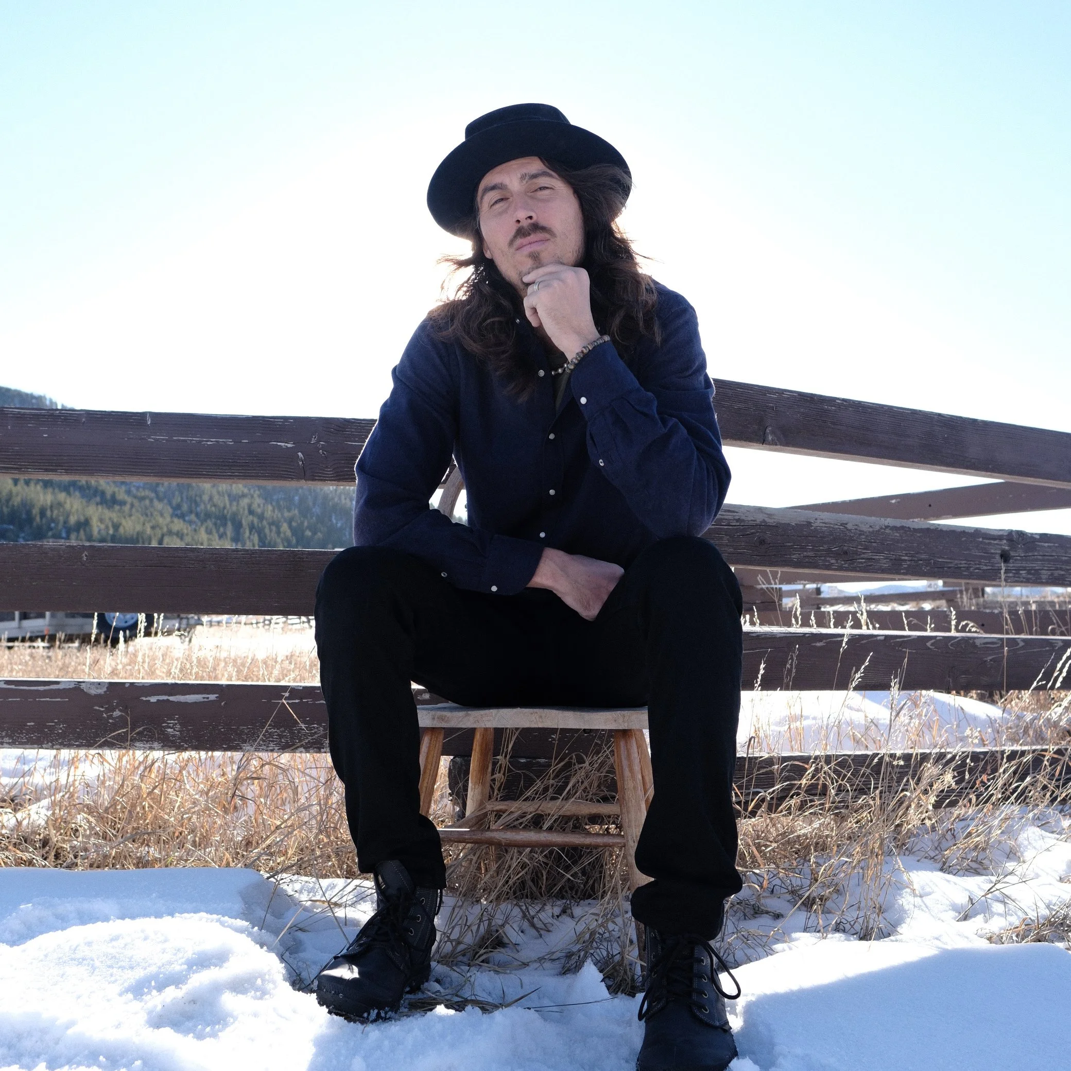 Dylan Townsend with long dark hair and a mustache, wearing a black hat and blue coat, sitting on a wooden bench in snowy outdoor setting, looking thoughtfully at the camera.