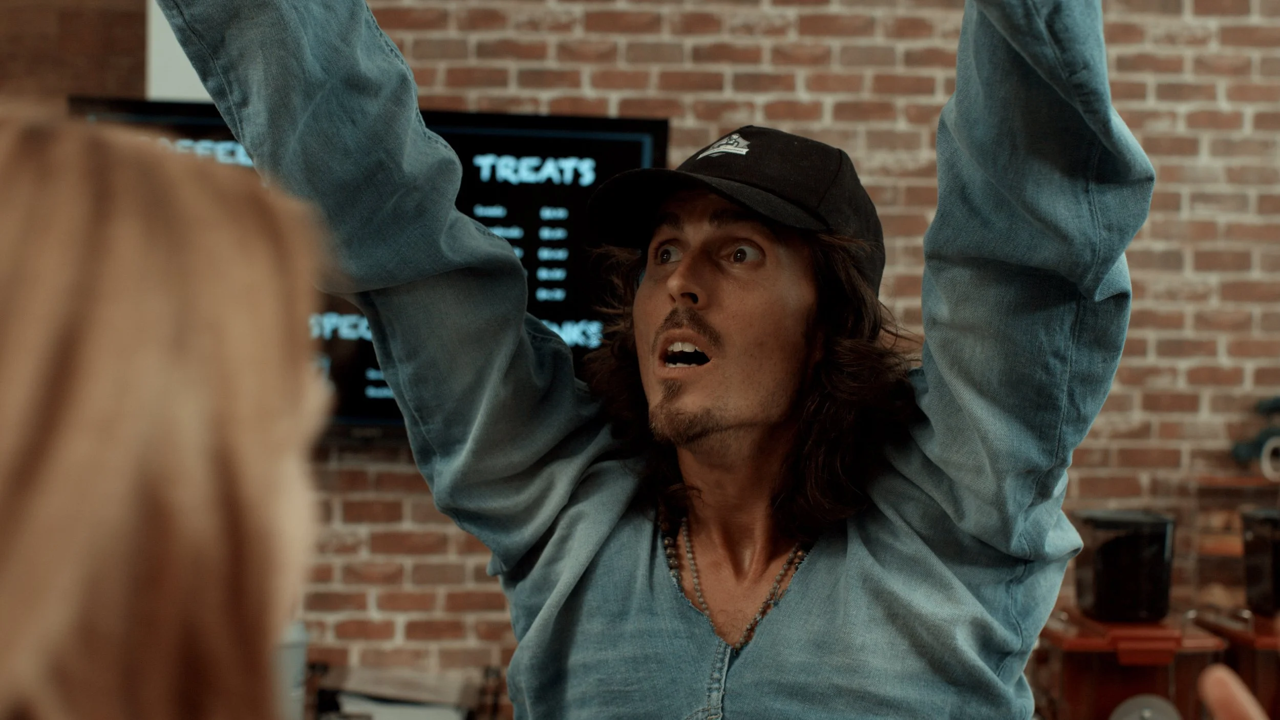 Dylan Townsend. Under the influence. with long hair wearing a black baseball cap and denim jacket pulling another person's hair, with an expression of surprise or fear, in front of a brick wall.