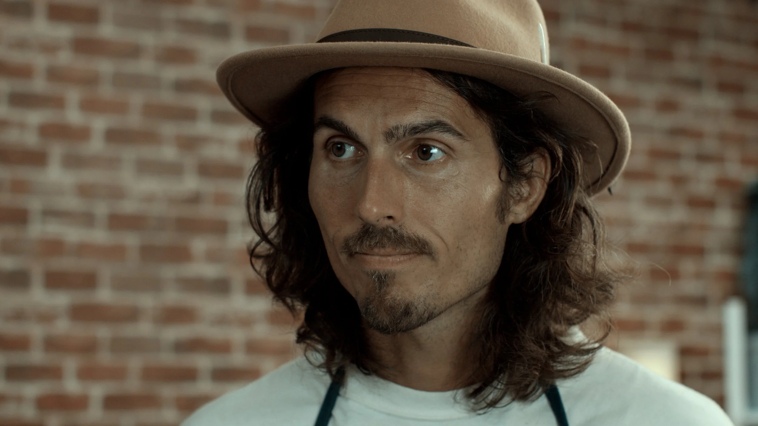 Dylan Townsend with long, curly brown hair, a beard, and mustache wearing a beige fedora hat and white shirt with a dark apron, standing in front of a brick wall.