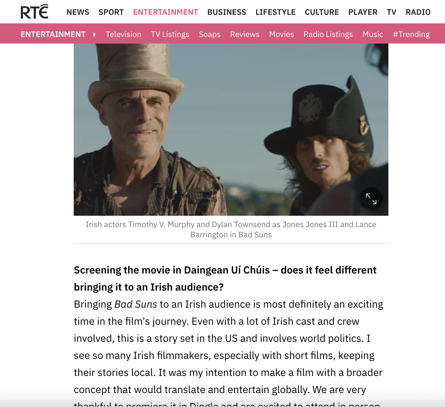Screenshot of a webpage from RTÉ entertainment section showing two actors dressed in period costumes with hats, standing outdoors with a clear sky in the background. The caption mentions Irish actors Timothy V. Murphy and Dylan Townsend as characters