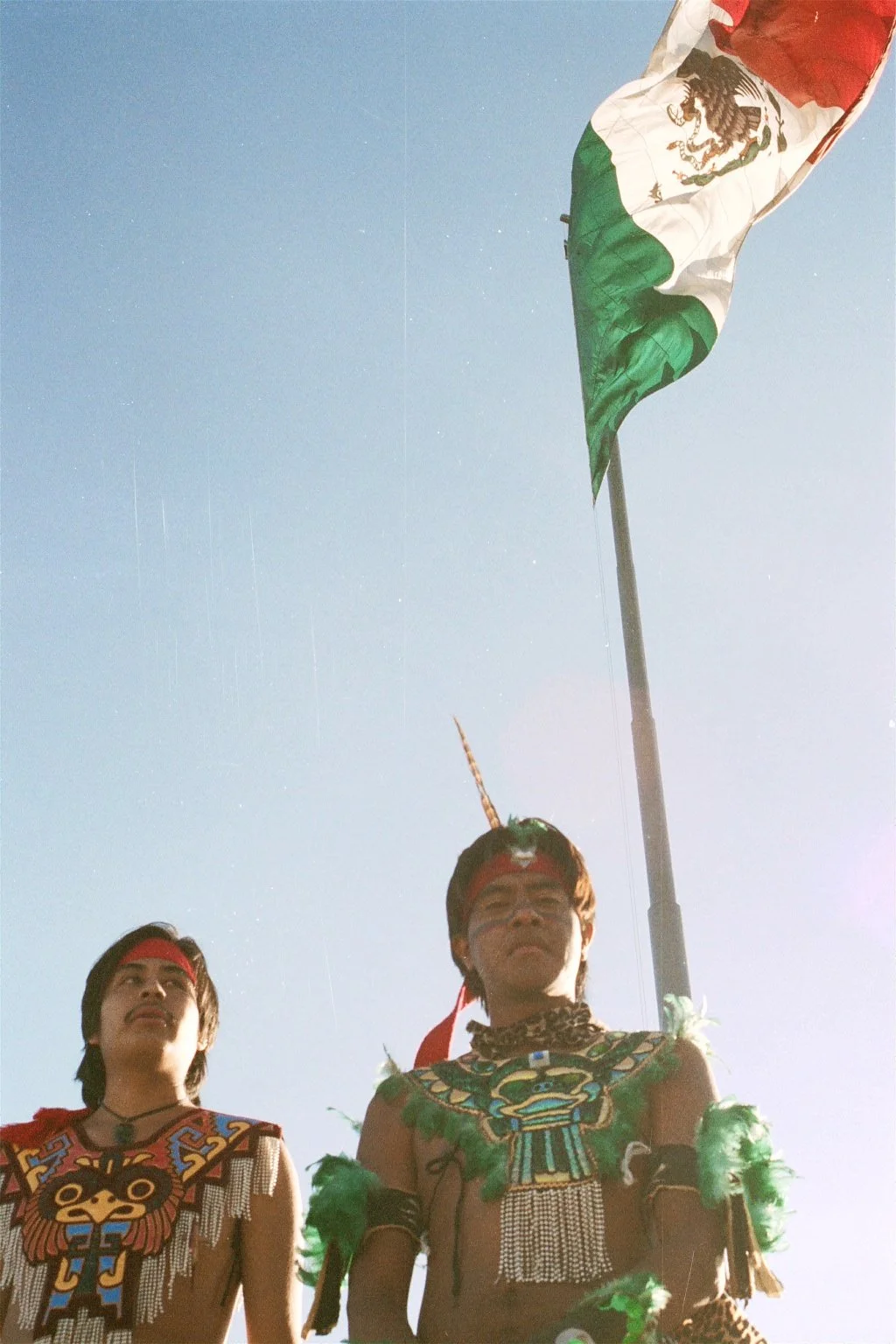 Two indigenous men wearing traditional attire with face paint, standing beneath a flag that appears to be the Mexican flag, against a clear blue sky.