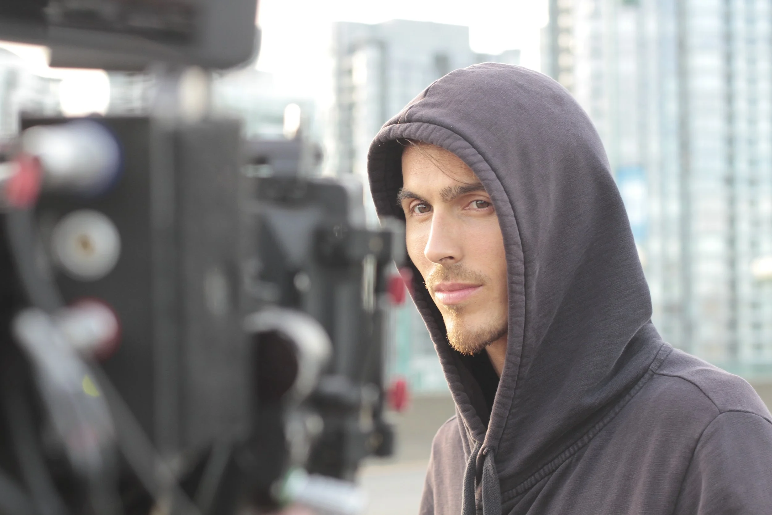 Dylan Townsend in film Heart Overheard. wearing a gray hoodie looks at a camera in an urban setting with modern glass buildings in the background.