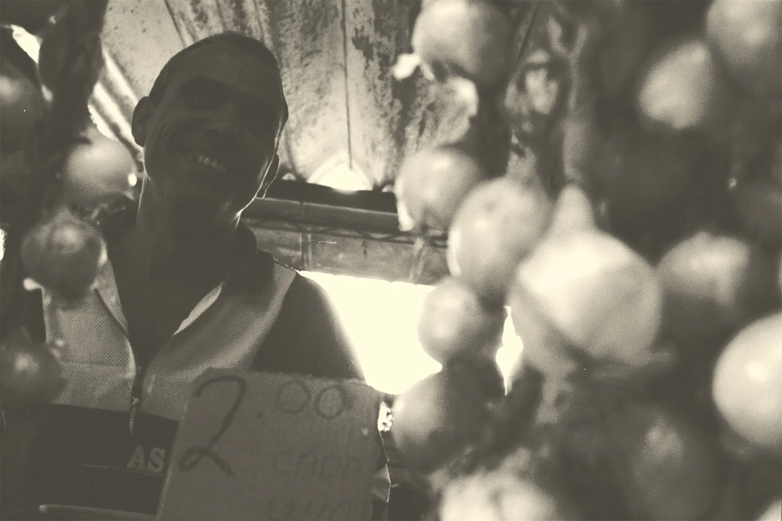 A person smiling, wearing glasses and a sleeveless top, standing behind a Christmas tree decorated with ornaments, with the background showing a rustic ceiling and a light source.