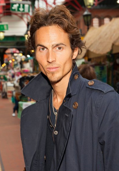 Young Dylan Townsend with curly brown hair and a serious expression, wearing a navy jacket outdoors in a market street with shops and people in the background.