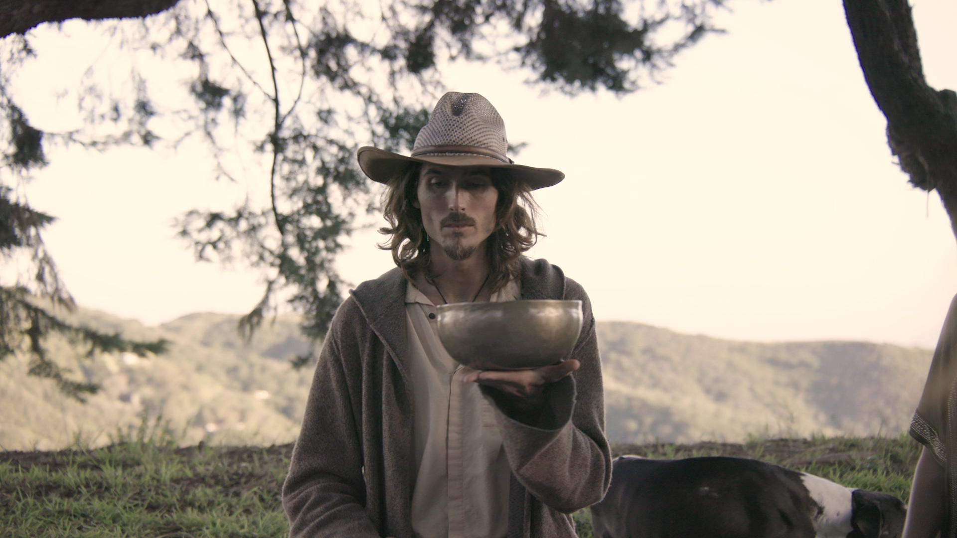 Dylan Townsend in Tv pilot 'Intentional' with shoulder-length hair and a beard wearing a wide-brimmed hat and a brown jacket is holding a metal bowl outdoors, with a scenic background of mountains, trees, and cloudy sky.