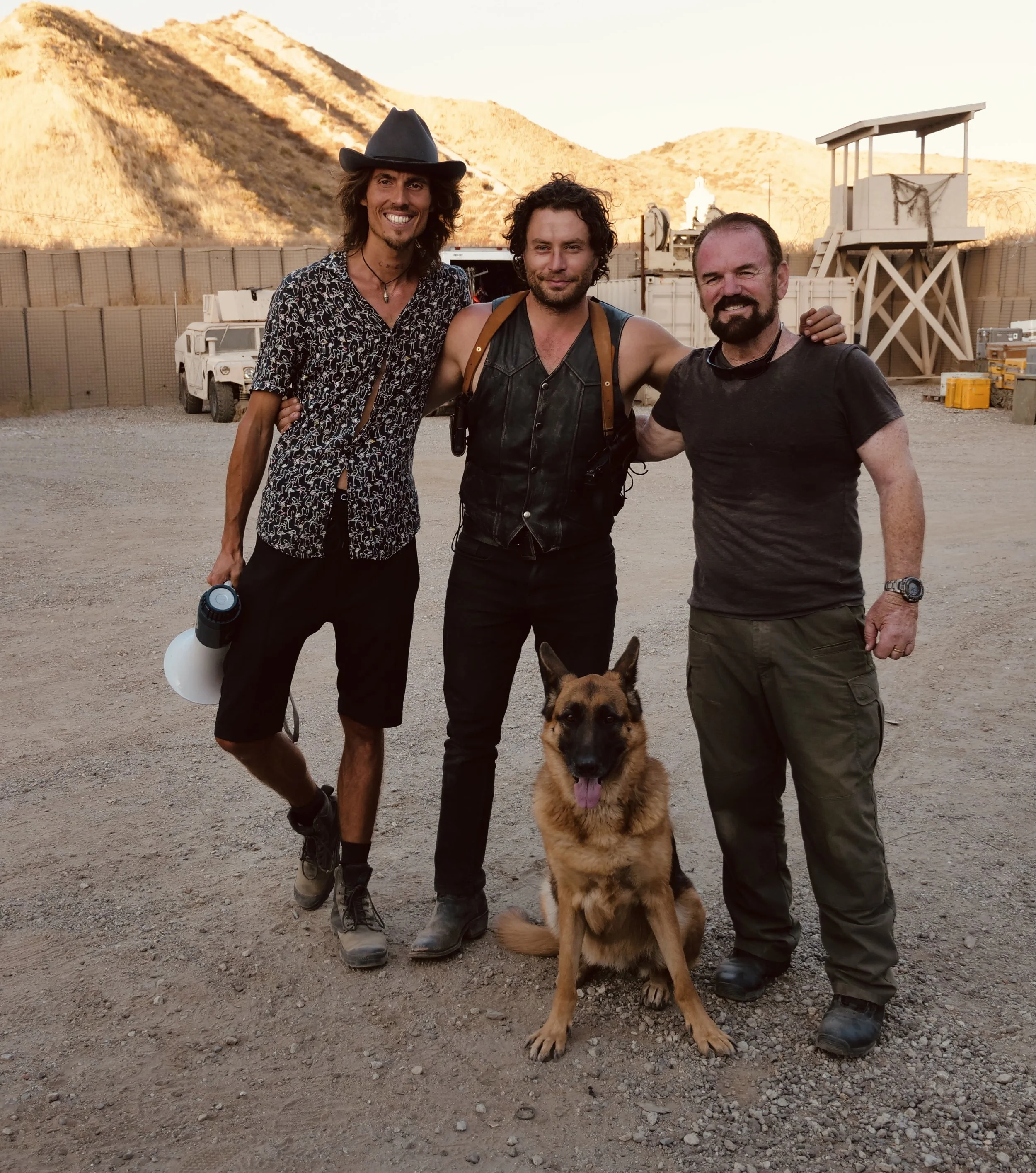 Dylan Townsend  & Kevin Ryan on set Film BAD SUNS. Three men and a German shepherd dog standing on a dirt ground outdoors with mountains and a watchtower in the background.