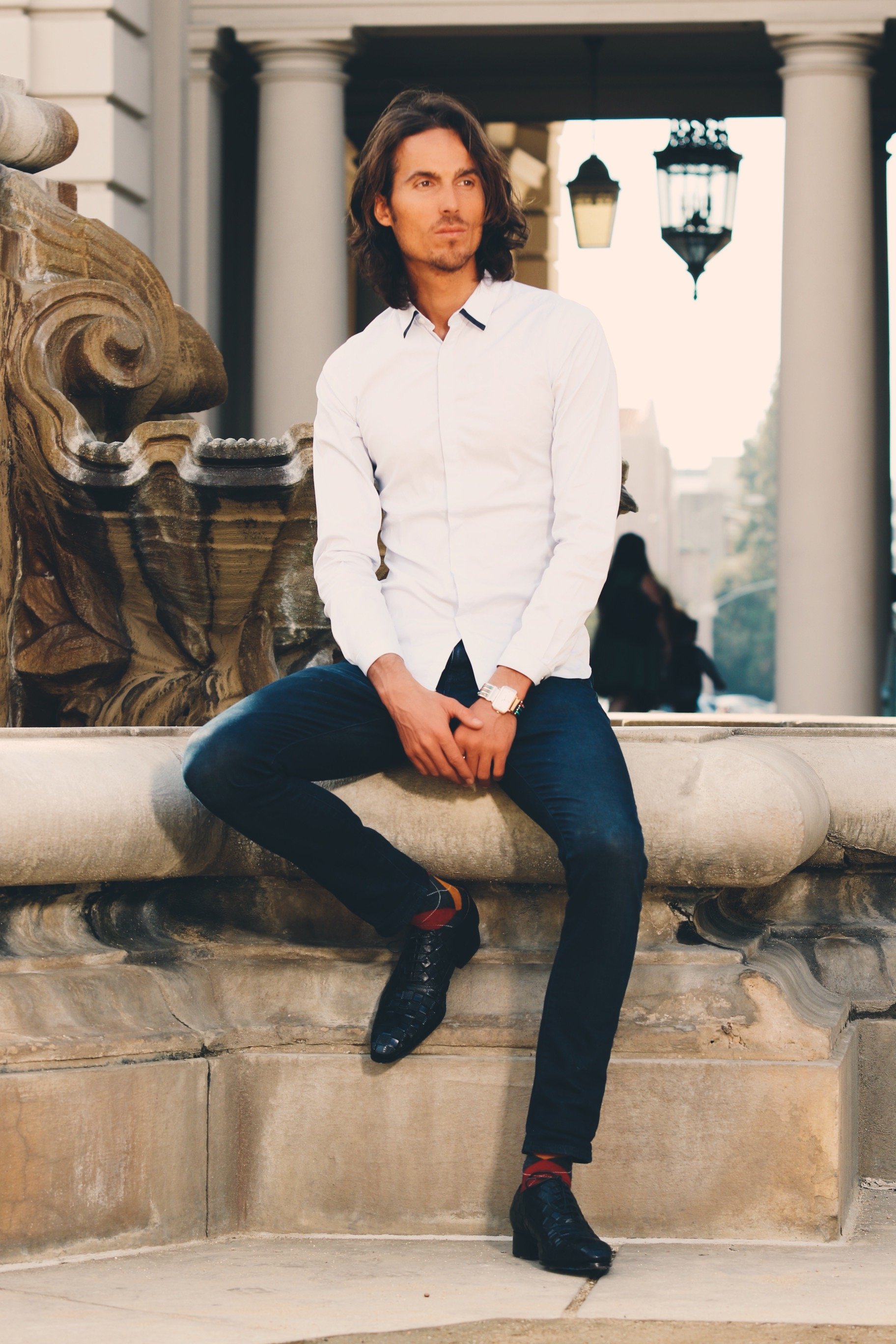 Dylan Townsend with long wavy dark hair and light skin sitting on a stone bench with an ornate face sculpture, wearing a white dress shirt, black pants, and black shoes, outdoors in front of a building with columns and hanging lanterns.