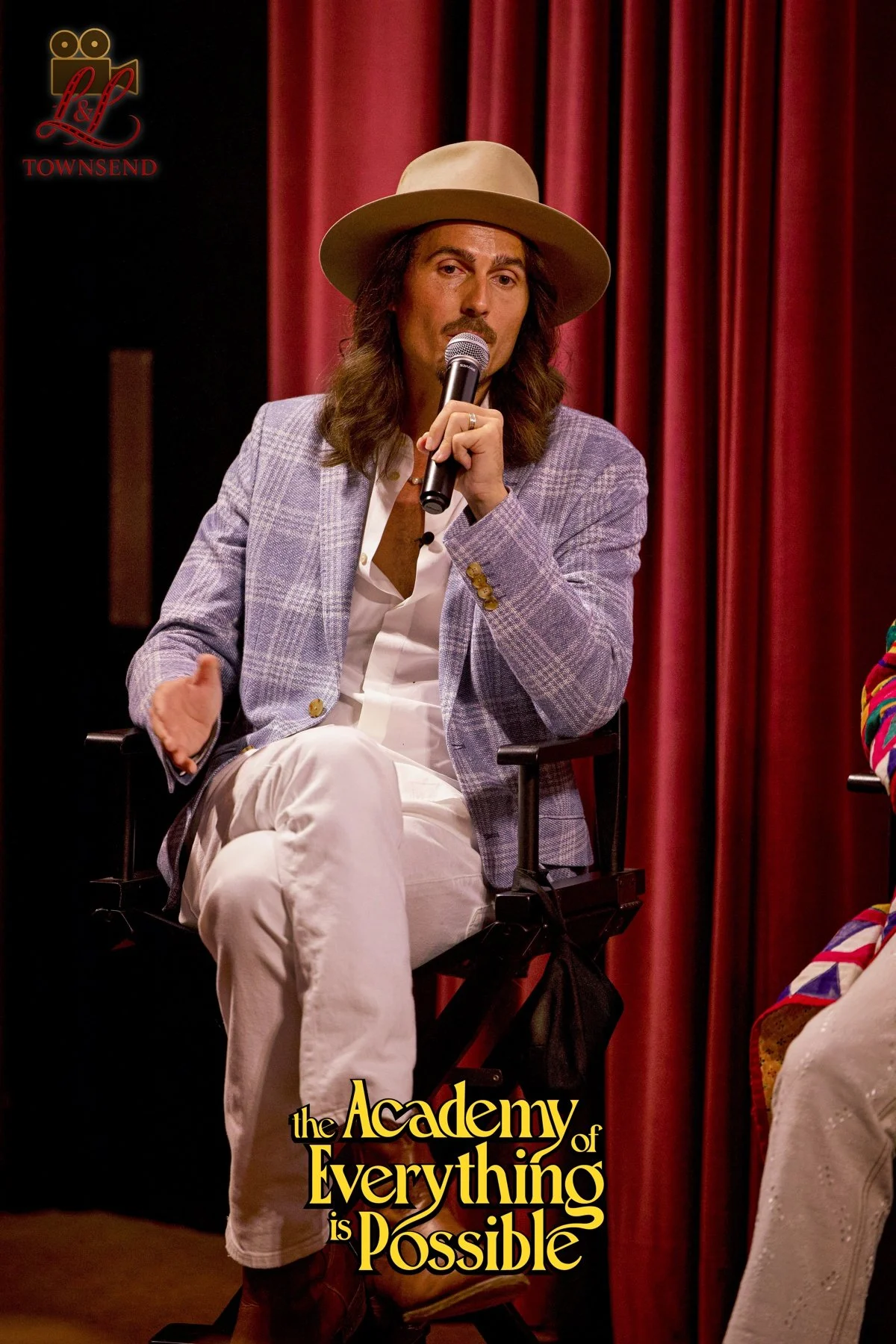 Dylan Townsend at private screening of feature film with long hair, a mustache, and beard, wearing a beige hat, a light purple plaid blazer, a white shirt, and beige pants, sitting on a chair on stage with red curtains behind him.