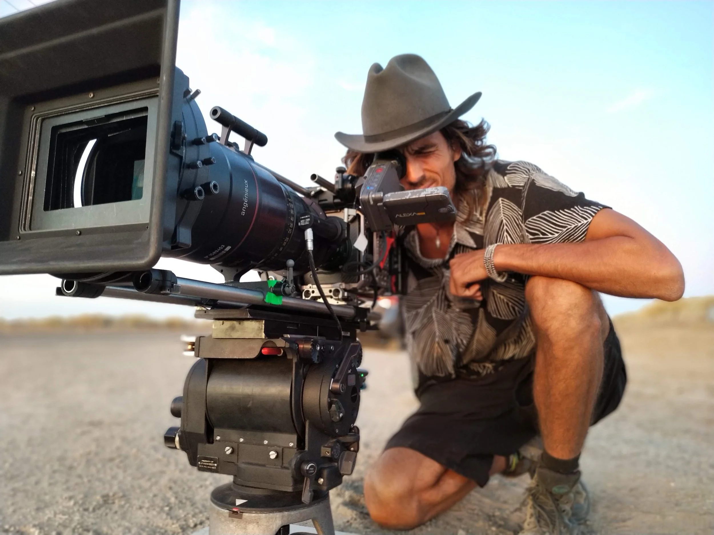 Dylan Townsend wearing a cowboy hat and patterned shirt using a professional camera mounted on a tripod outdoors during daytime.