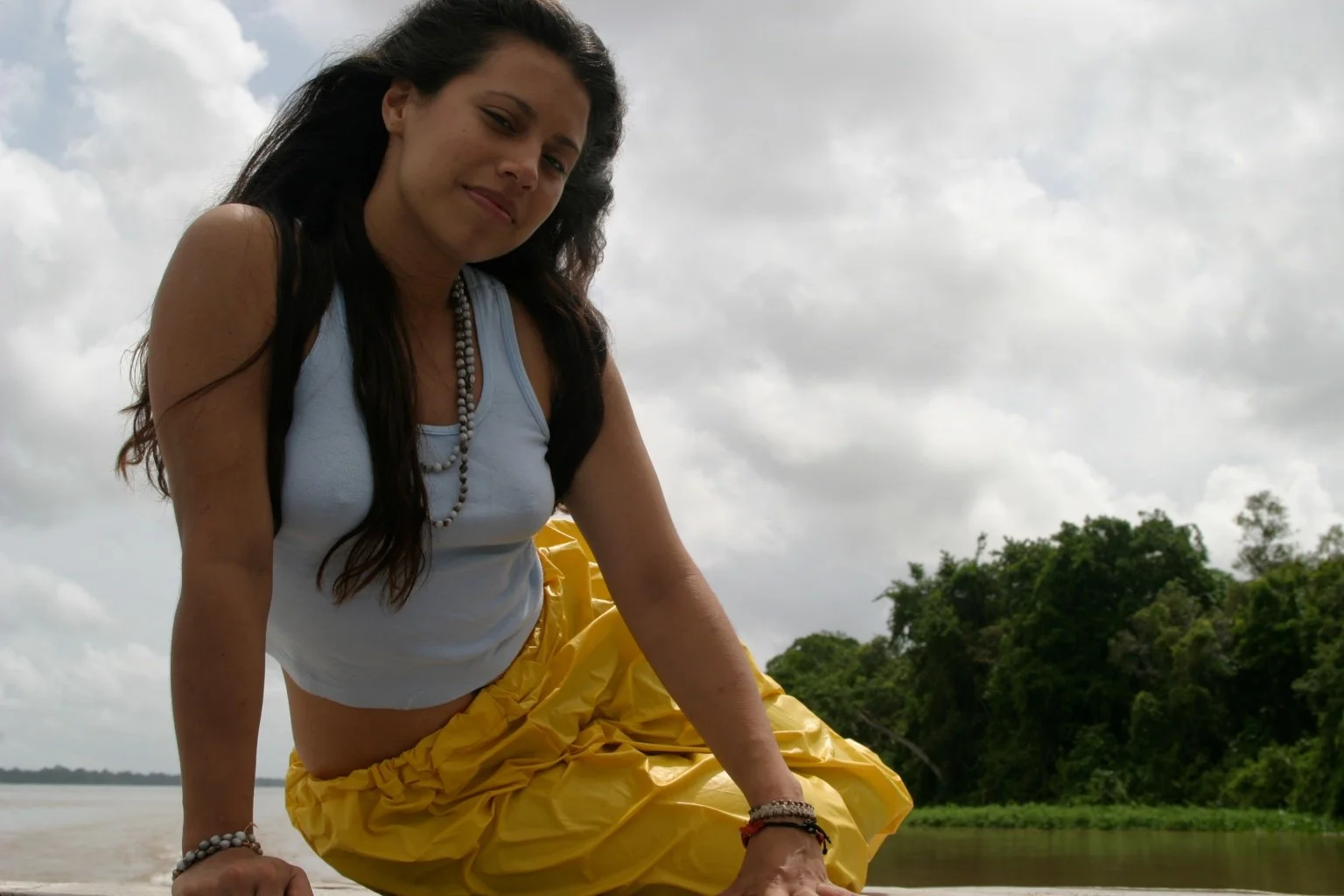 A woman with long dark hair wearing a white tank top, yellow pants, and jewelry, sitting outdoors near a river and trees with an overcast sky in the background.