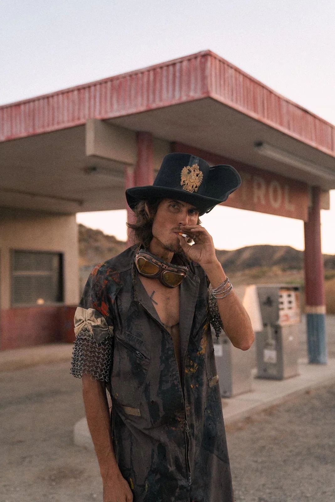 Dylan Townsend with long hair and a mustache, wearing a large cowboy hat with a badge, smoking a cigarette, standing outside an old building with a pink roof.