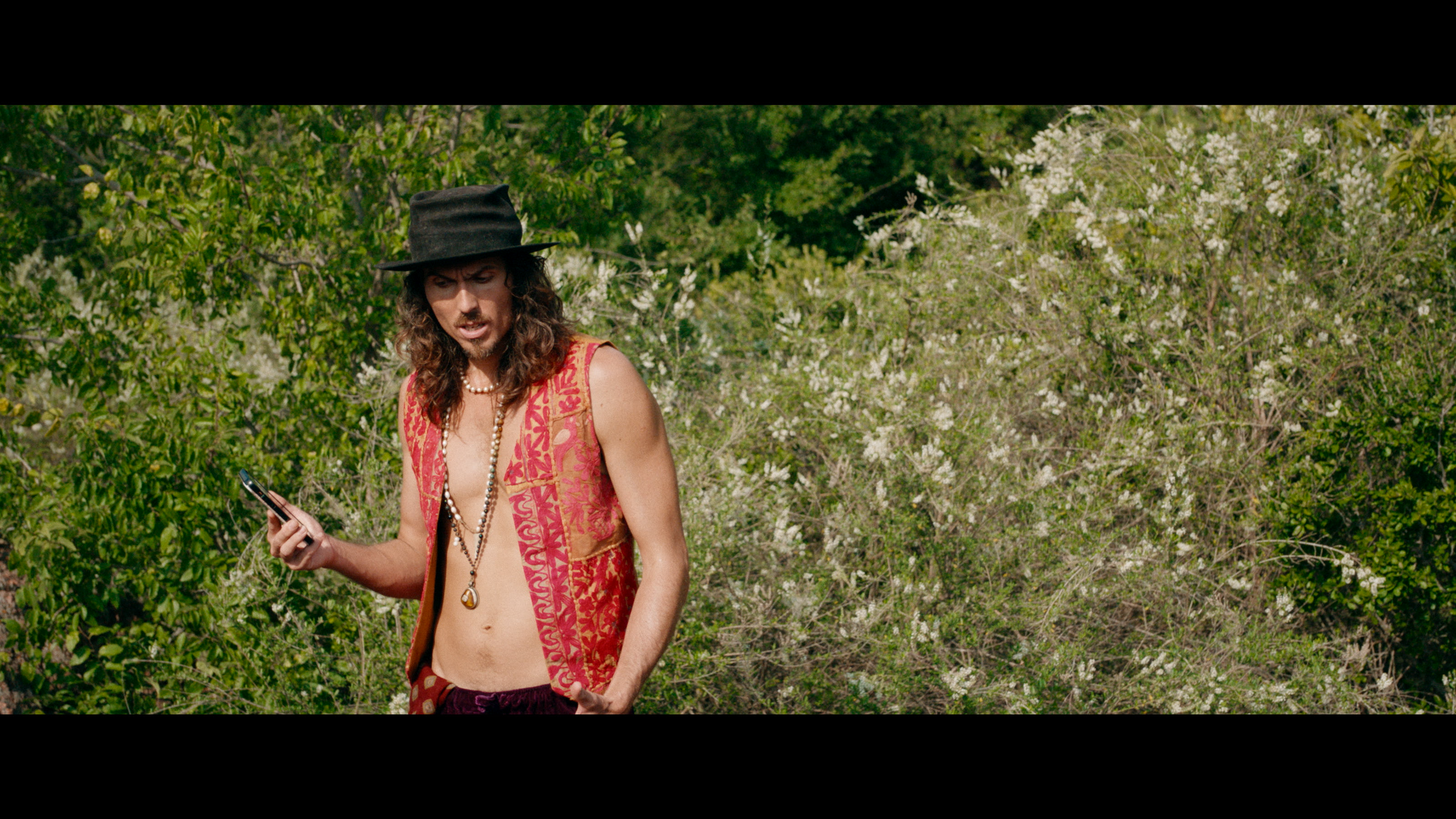 Dylan Townsend with long curly hair and a hat wearing an open vest and necklaces, looking at his phone outdoors with green trees and bushes in the background.