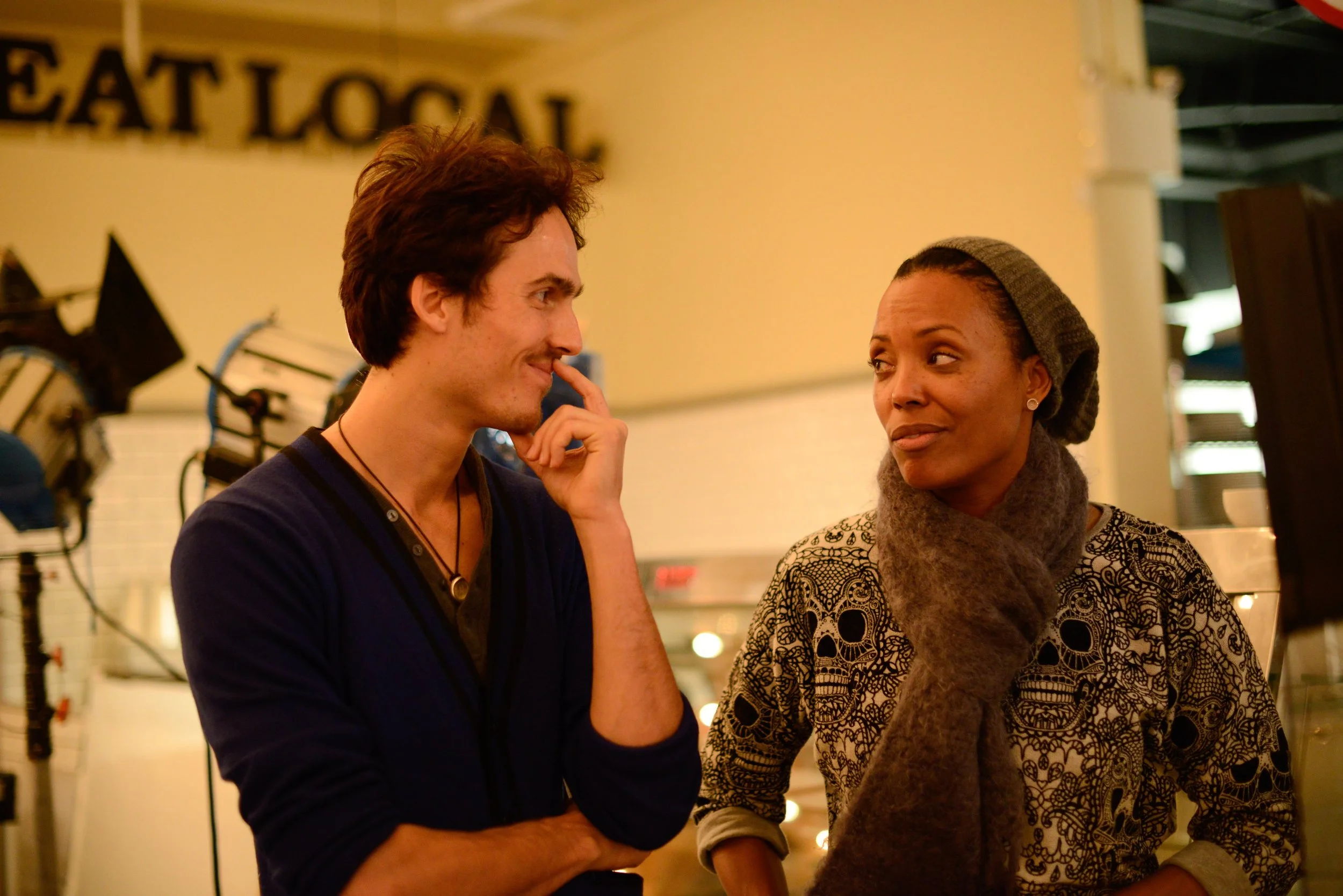 Dylan Townsend & Aisha Tyler engaging in conversation indoors, with the man touching his face and smiling and the woman looking at him with a slight smile. The background includes a sign partially reading 'EAT LOCAL' and various blurred objects.