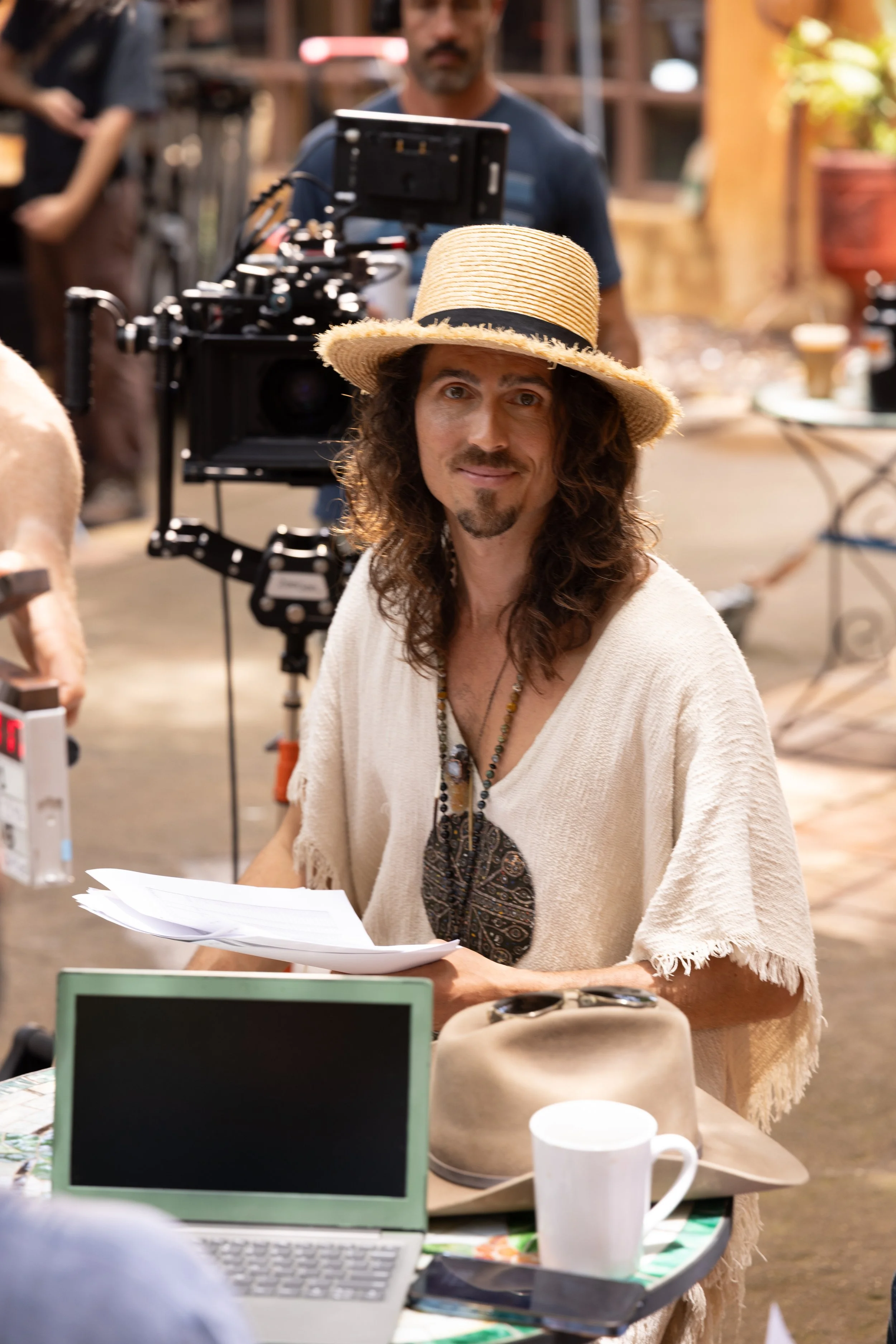 Dylan Townsend on set The Academy of Everything is Possible with long curly hair, a mustache, and a goatee wearing a straw hat and a beige poncho, sitting at a table with papers, with film crew and camera equipment in the background.