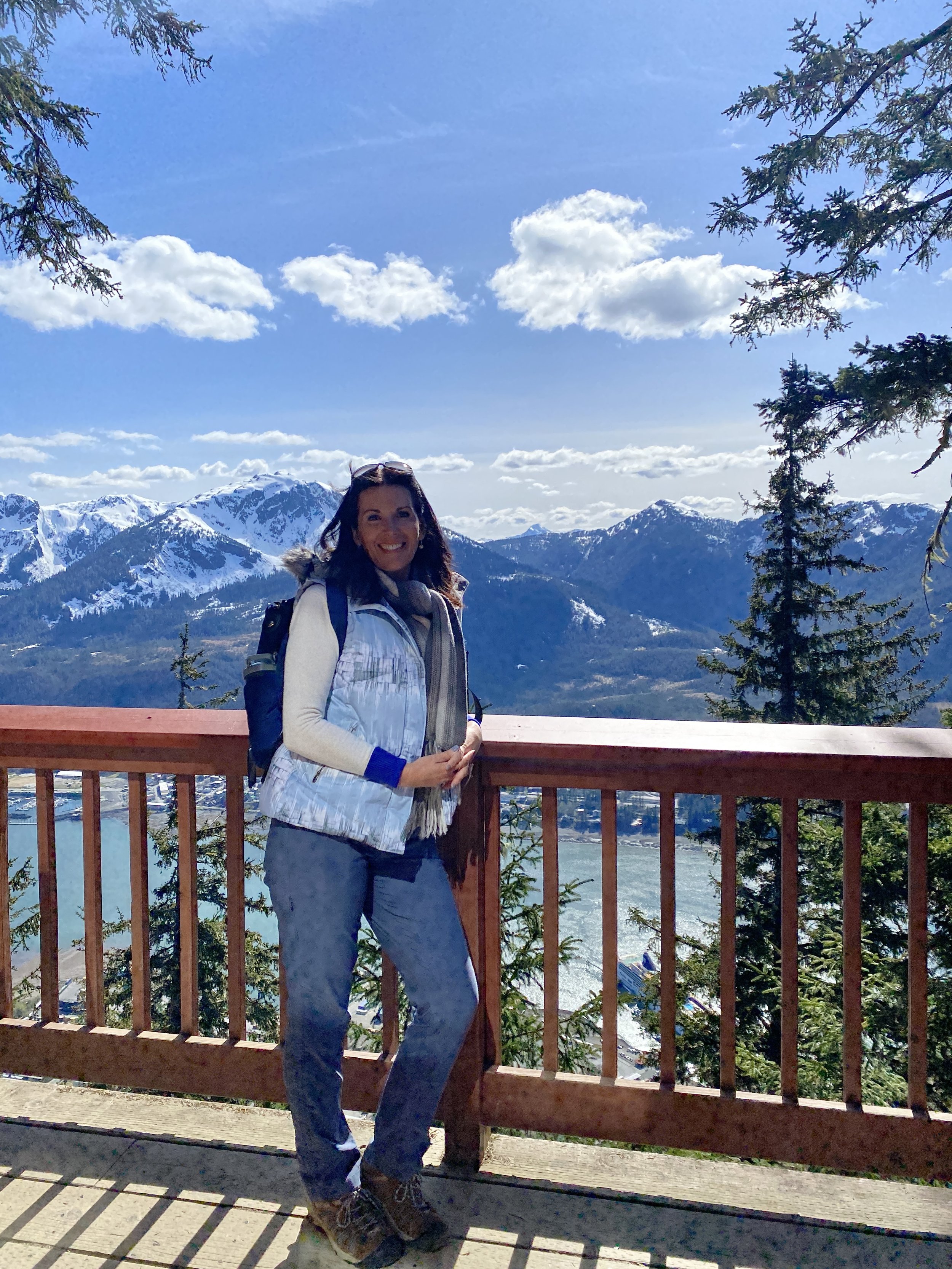 A woman standing on a balcony or viewing platform with a mountain range with snow-capped peaks in the background. She is smiling, wearing outdoor clothing and sunglasses on her head, and has a backpack. The sky is blue with some scattered clouds and evergreen trees frame the scene.
