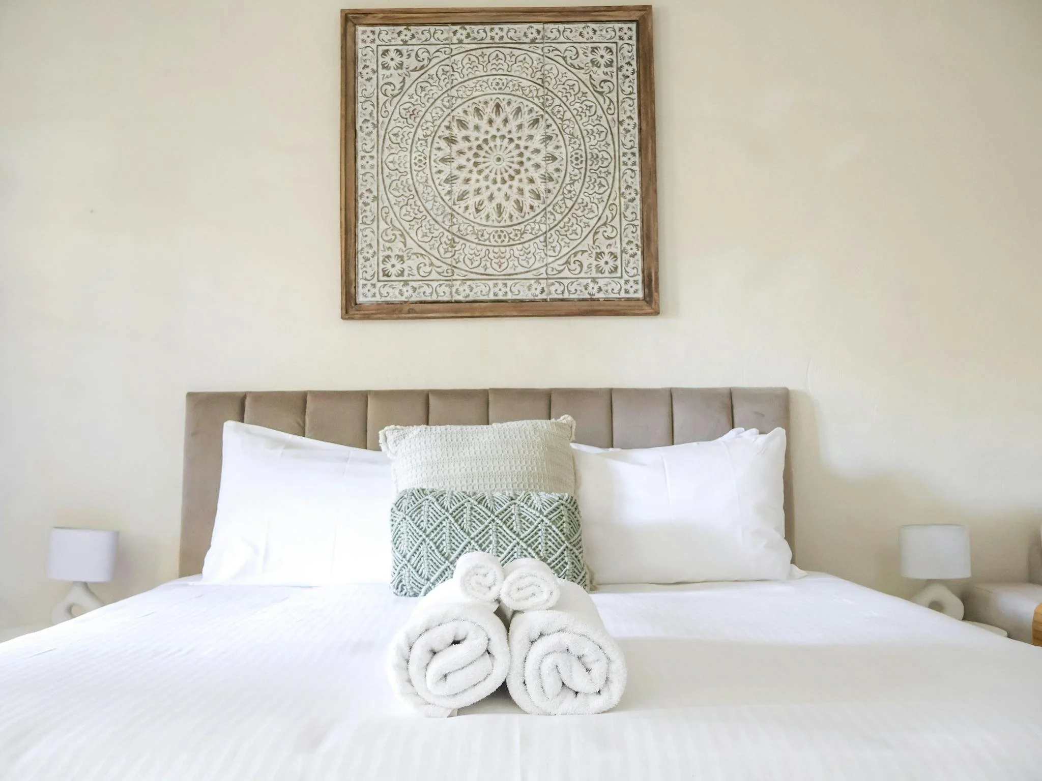 A neatly made bed with white bedding, a pale beige upholstered headboard, and decorative pillows. Four rolled white towels are placed at the foot of the bed. Above the bed, there is a framed wall art piece with a circular, intricate carved design. Small white lamps are visible on each side of the bed.
