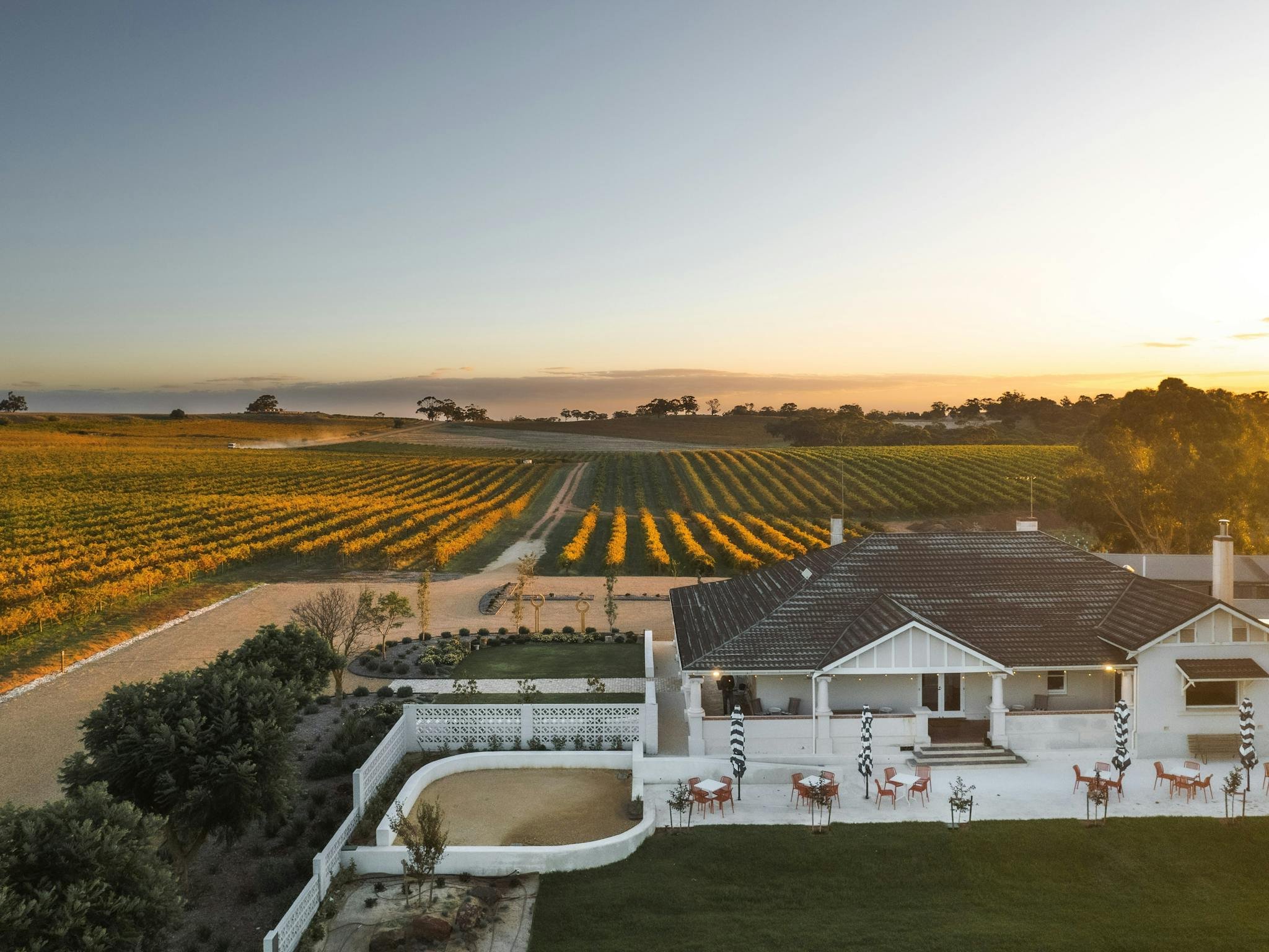A house with a large backyard patio, white fencing, and outdoor furniture, overlooking a scenic agricultural landscape with fields and trees during sunset.