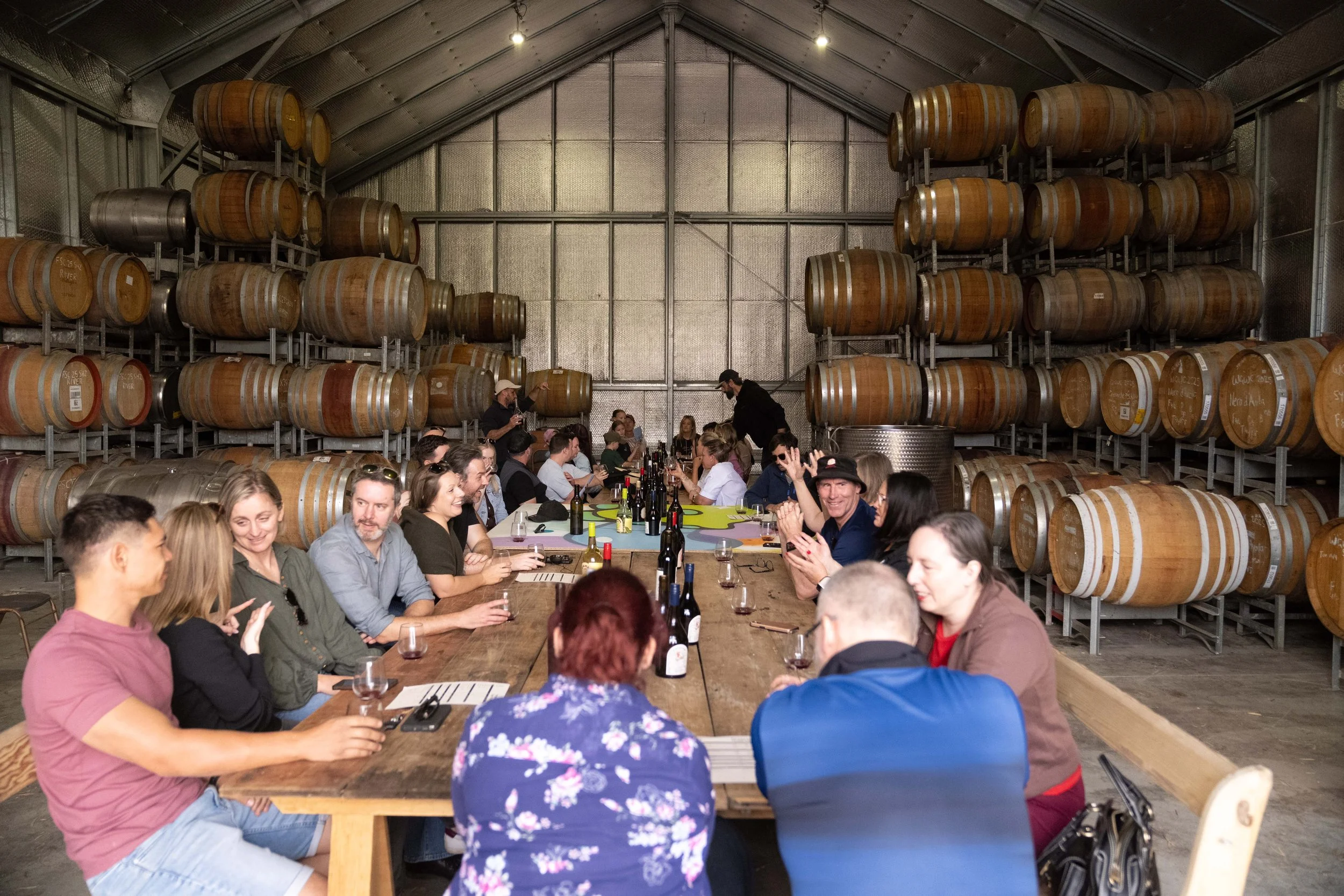 A group of people sitting around a long wooden table inside a winery, with rows of wine barrels stacked on metal racks against the walls in the background.