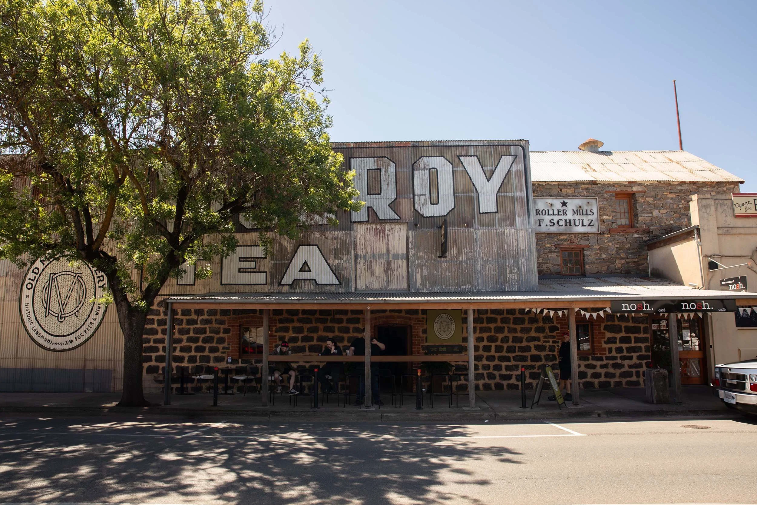 Exterior of a rustic building with a large sign that reads 'RUY' and smaller signs advertising roller mills and a cafe. There are trees in the foreground, and a few people are sitting at a shaded outdoor seating area.