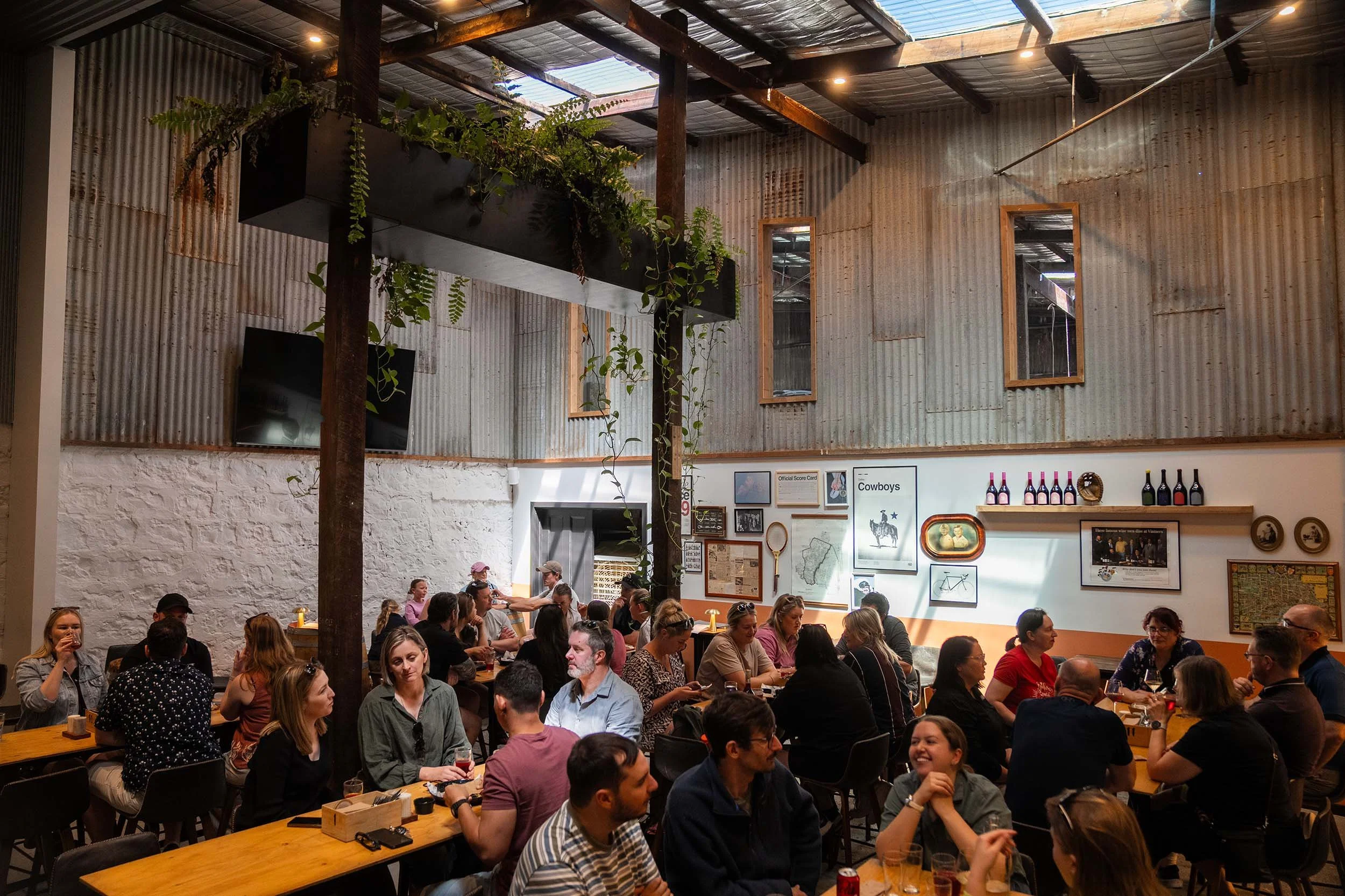 Crowd of people dining in an indoor restaurant with rustic corrugated metal walls, wooden beams, and overhead skylights, with some framed pictures and bottles on a shelf.