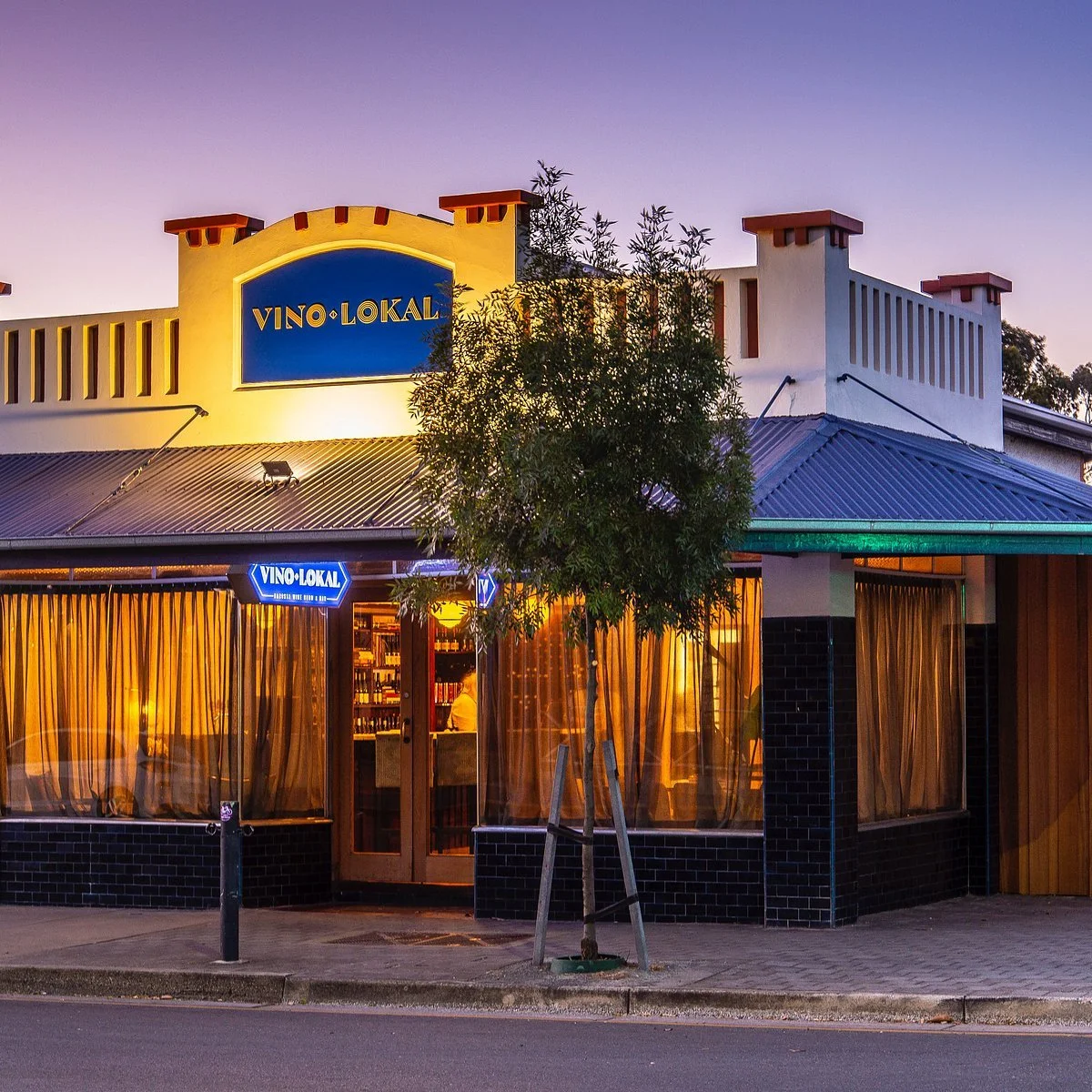 Exterior view of a restaurant called 'Vino Lokal' during twilight, with a tree in front and large windows revealing the interior.