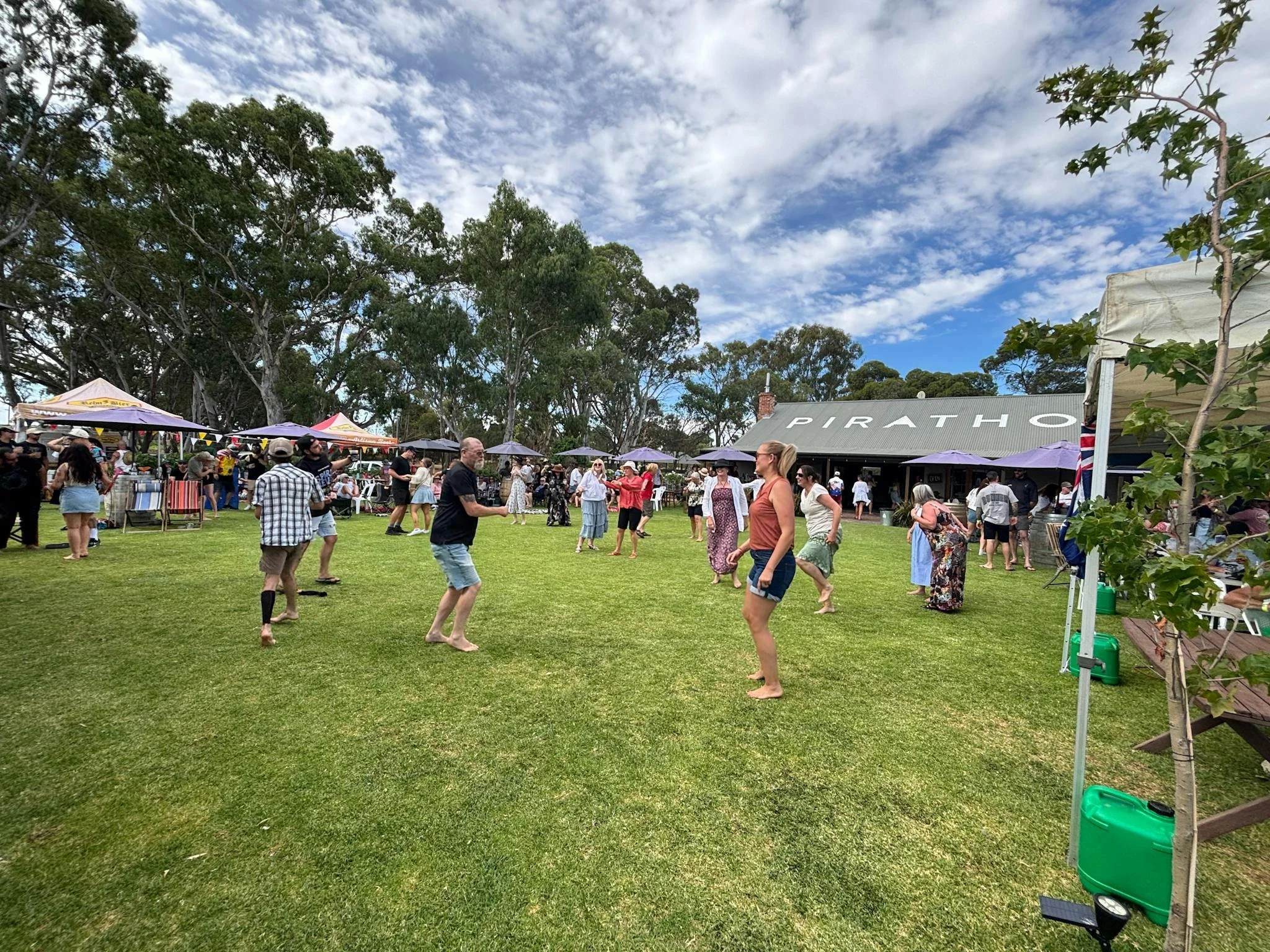 People gathering outdoors at a daytime event with tents and a building labeled 'PIRATHO', large trees, a cloudy sky, and green grass.