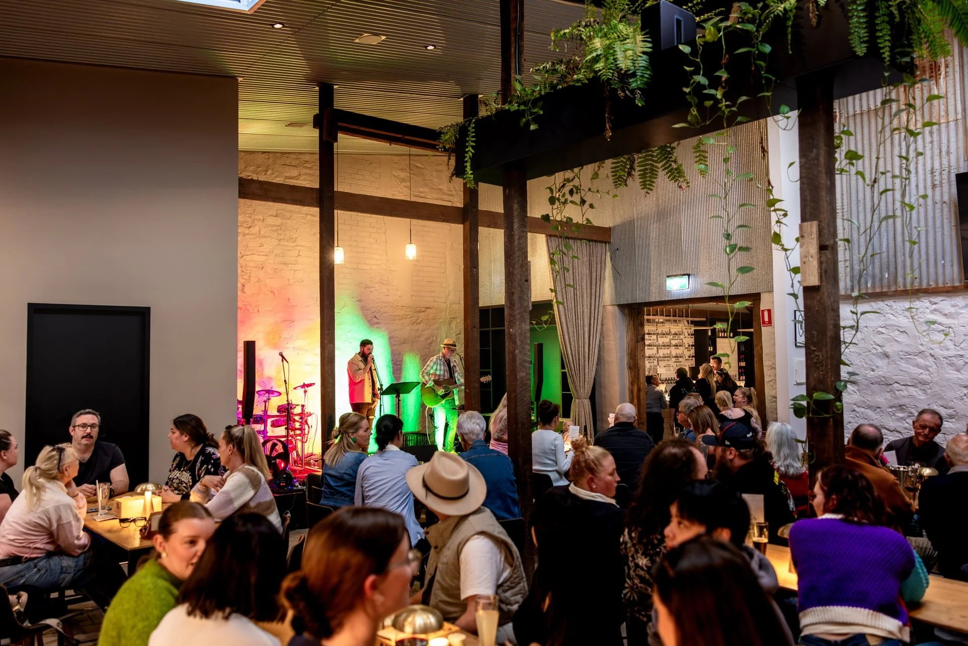 A lively indoor concert with a musical band performing on stage and an audience in a restaurant or cafe setting, decorated with plants, hanging lights, and rustic interior design.