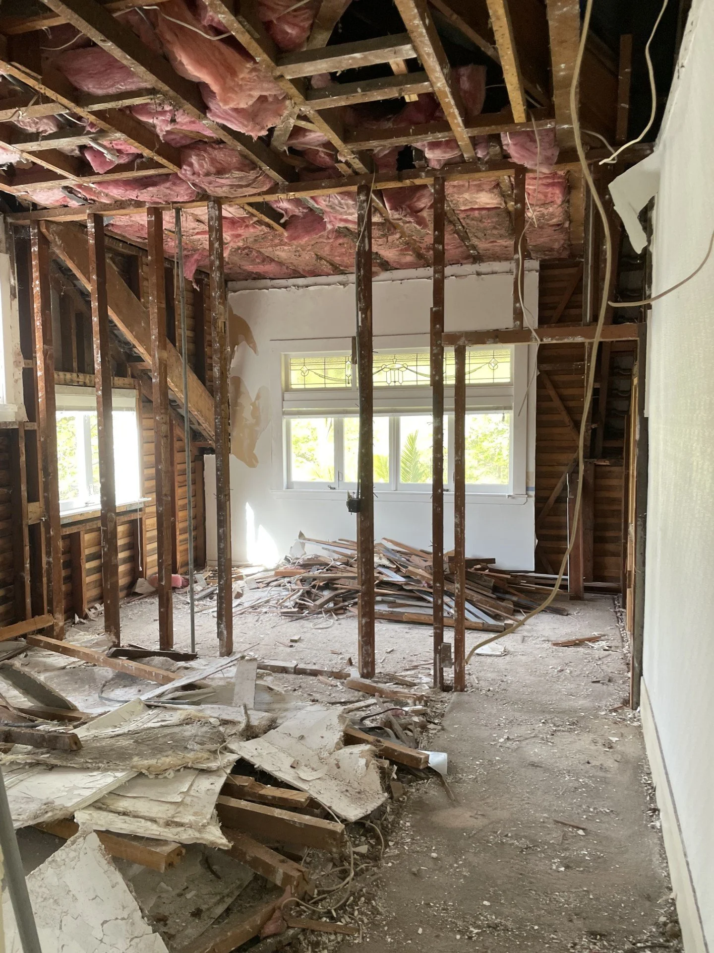 Interior of a house under renovation with exposed wall studs, pink insulation, debris on the floor, and a large window with sunlight streaming in.